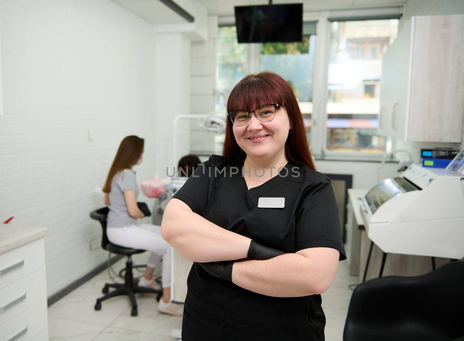 Caucasian positive committed young female dentist doctor looking at camera, standing with arms folded in dental office by artgf