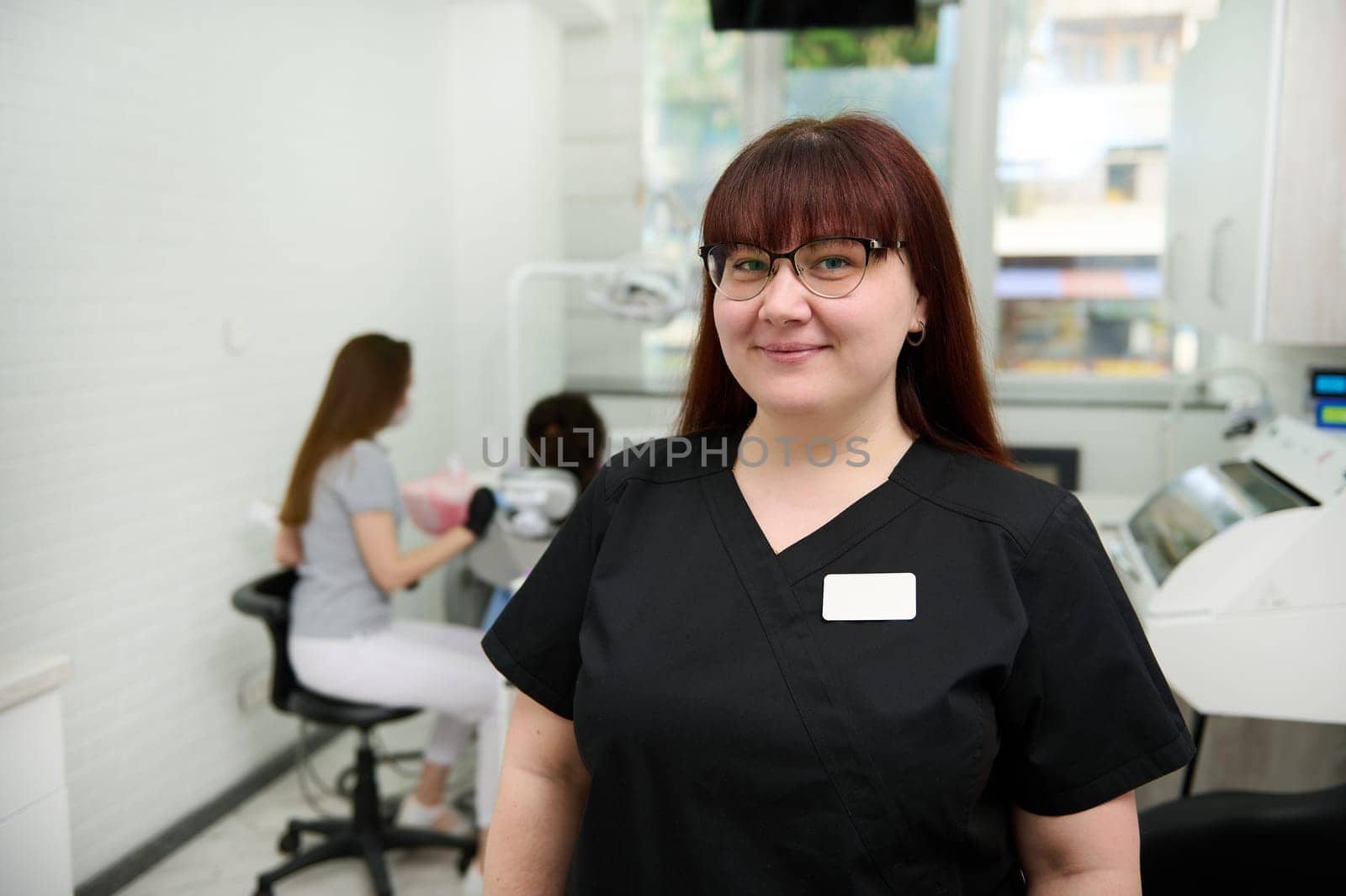 Confident portrait of Caucasian young positive happy female dentist doctor, orthodontist in black medical uniform, smiling looking at camera, standing in the dental office of a modern dentistry clinic