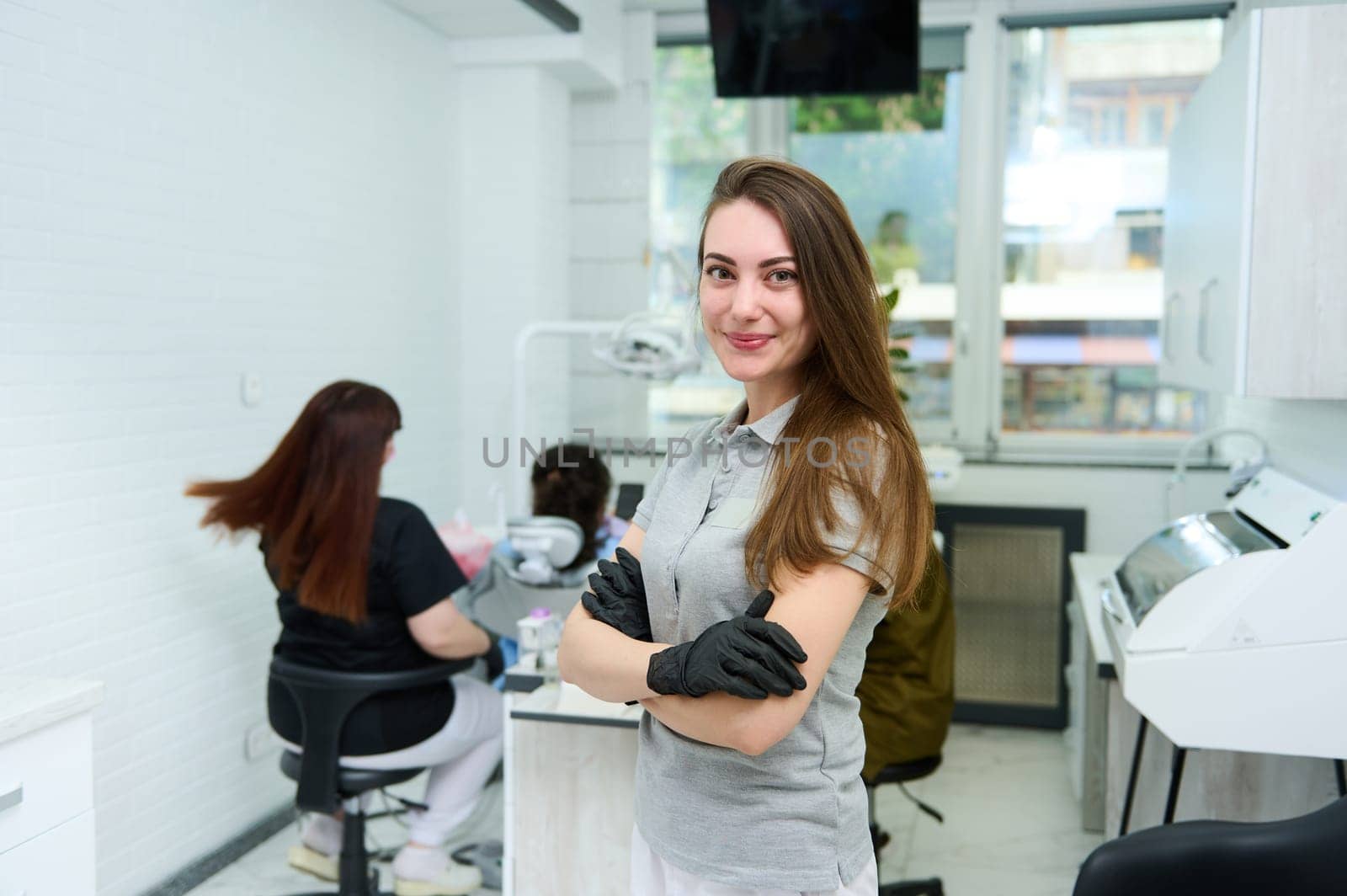 Caucasian charming woman 25-20 years old, wearing gray medical uniform, confident female dentist doctor, orthodontist, maxillofacial surgeon smiles at camera, stands with folded arms in dental clinic