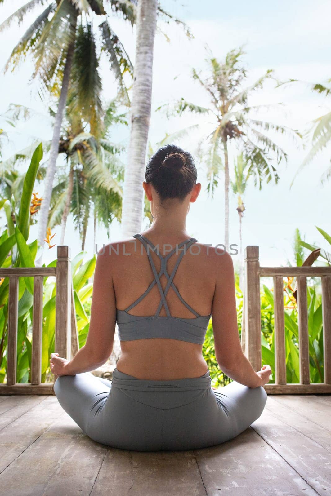 Young woman doing meditation in wooden porch in vacation summer resort. Rear view of young female meditating outdoors. Vertical image. Healthy lifestyle and mental health concept.