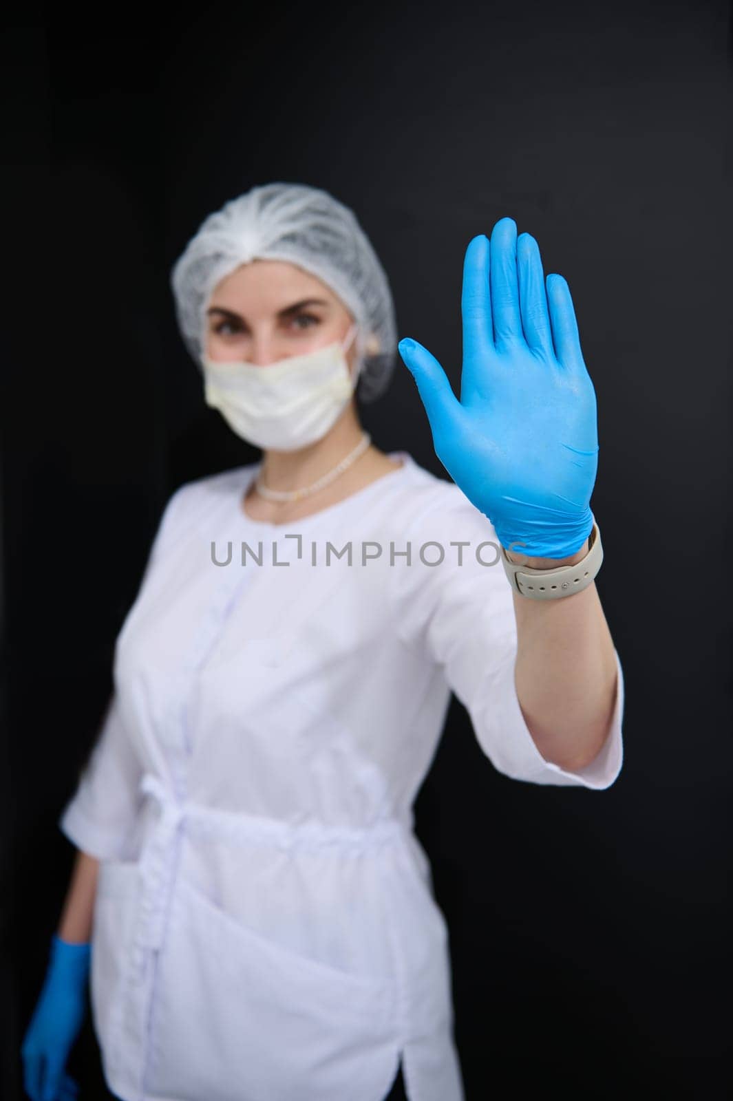 Focus on hand in blue protective latex glove, of blurred woman doctor, surgeon, medical staff, healthcare worker in white lab coat and protective medical mask, shows her hand palm to camera. STOP sign