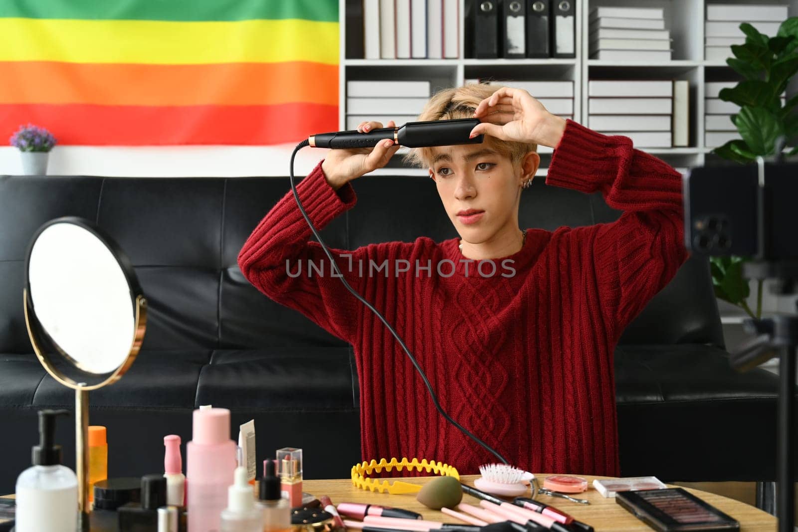 Attractive young gay man in red sweater straightening hair with flat iron. LGBTQ, domestic life, fashion and beauty concept.