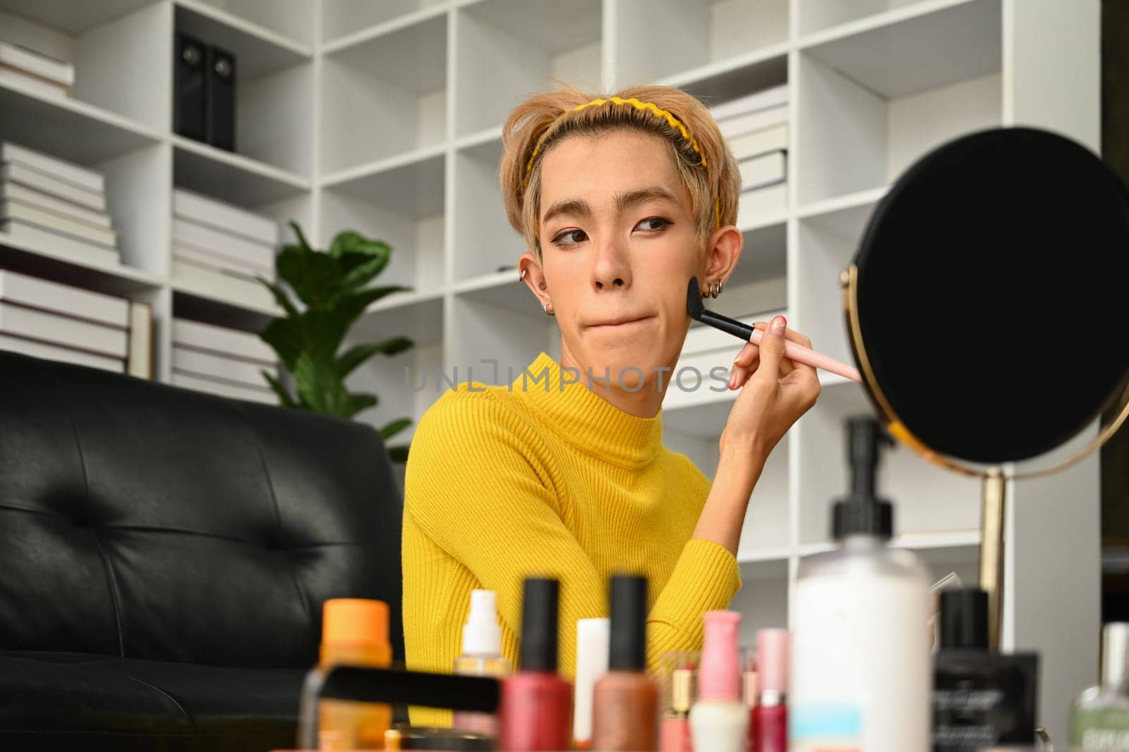 Young gay man doing daily makeup in living room, applying base foundation on face with sponge. LGBTQ, lifestyle and makeup concept.