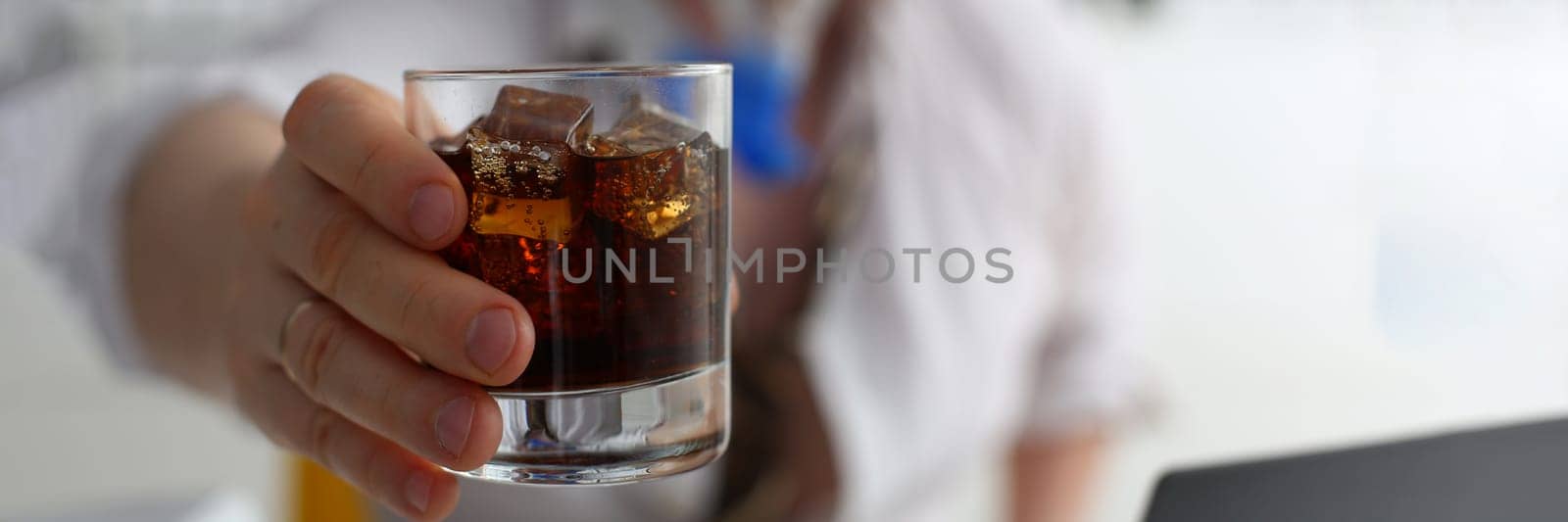 man holds a glass of cola and whiskey with ice in hand at workplace in office by kuprevich