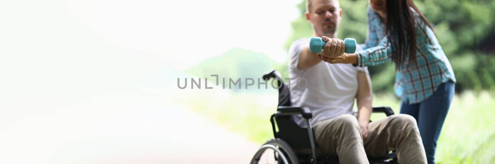 Female physiotherapist helps disabled man in wheelchair to lift weight of arm in park. Rehabilitation and exercises with dumbbells concept