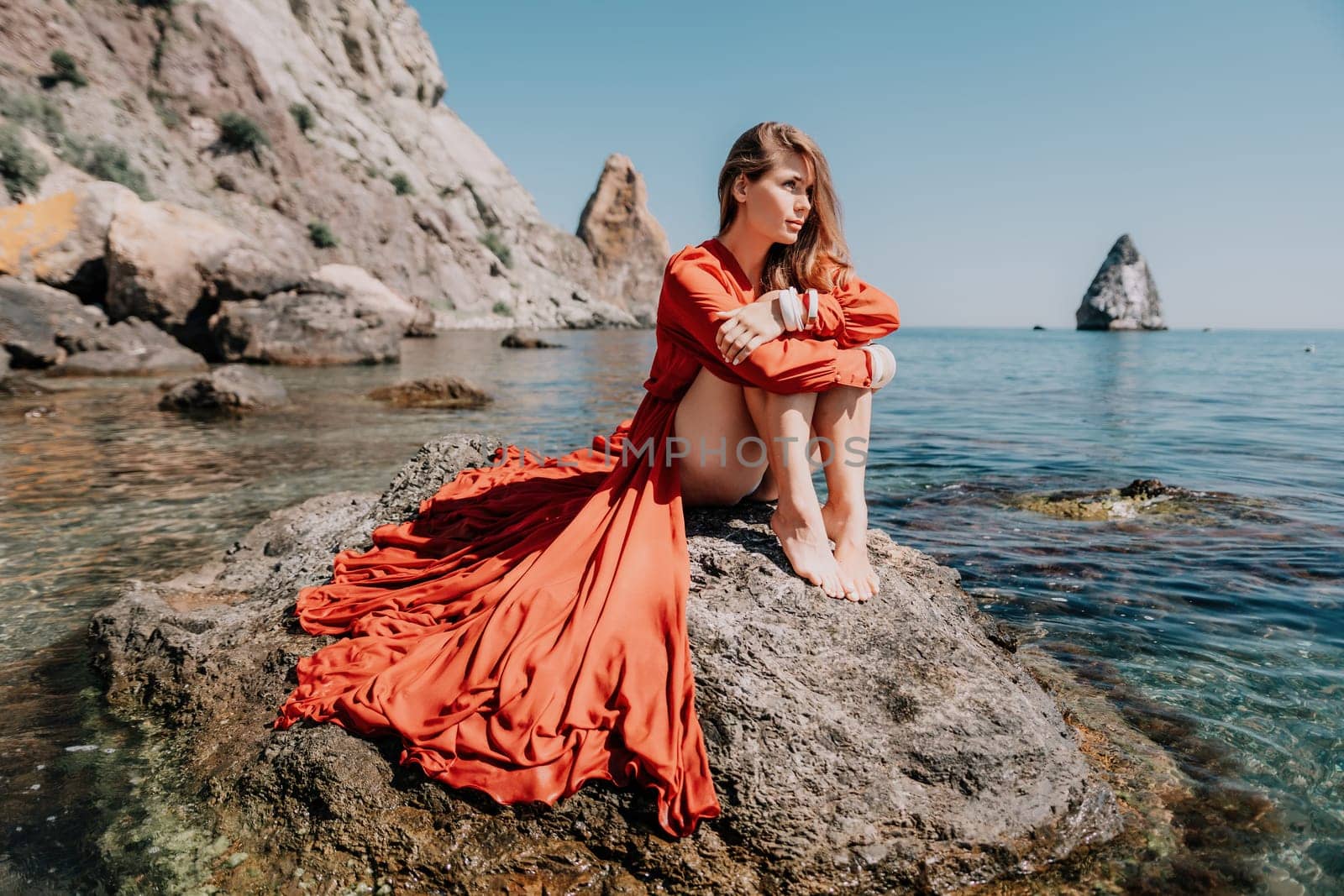 Woman travel sea. Happy tourist in red dress enjoy taking picture outdoors for memories. Woman traveler posing on the rock at sea bay surrounded by volcanic mountains, sharing travel adventure journey by panophotograph