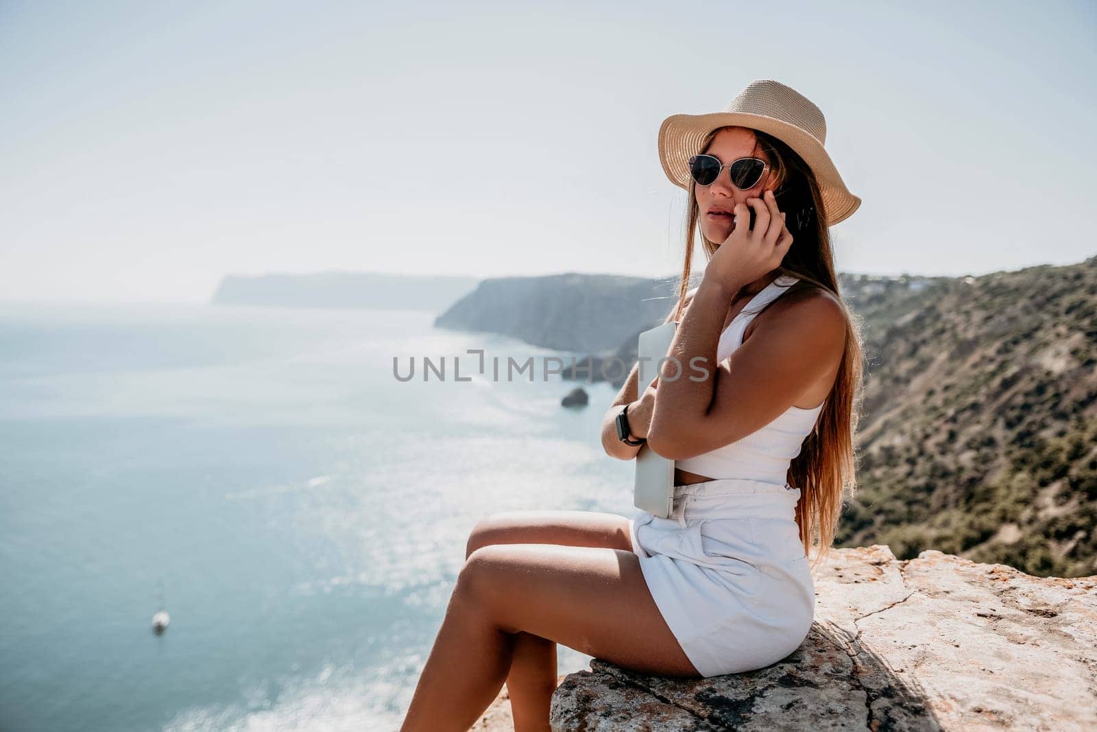 Successful business woman in yellow hat working on laptop by the sea. Pretty lady typing on computer at summer day outdoors. Freelance, travel and holidays concept.