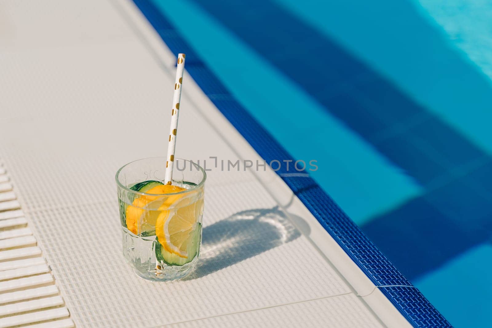 Tropical sparkling cocktail by the pool. The picture of glass with lemon and cucumber lemonade fruit cocktail standing near the poolside. Summer alcohol free drink. Hello summer holiday vacation