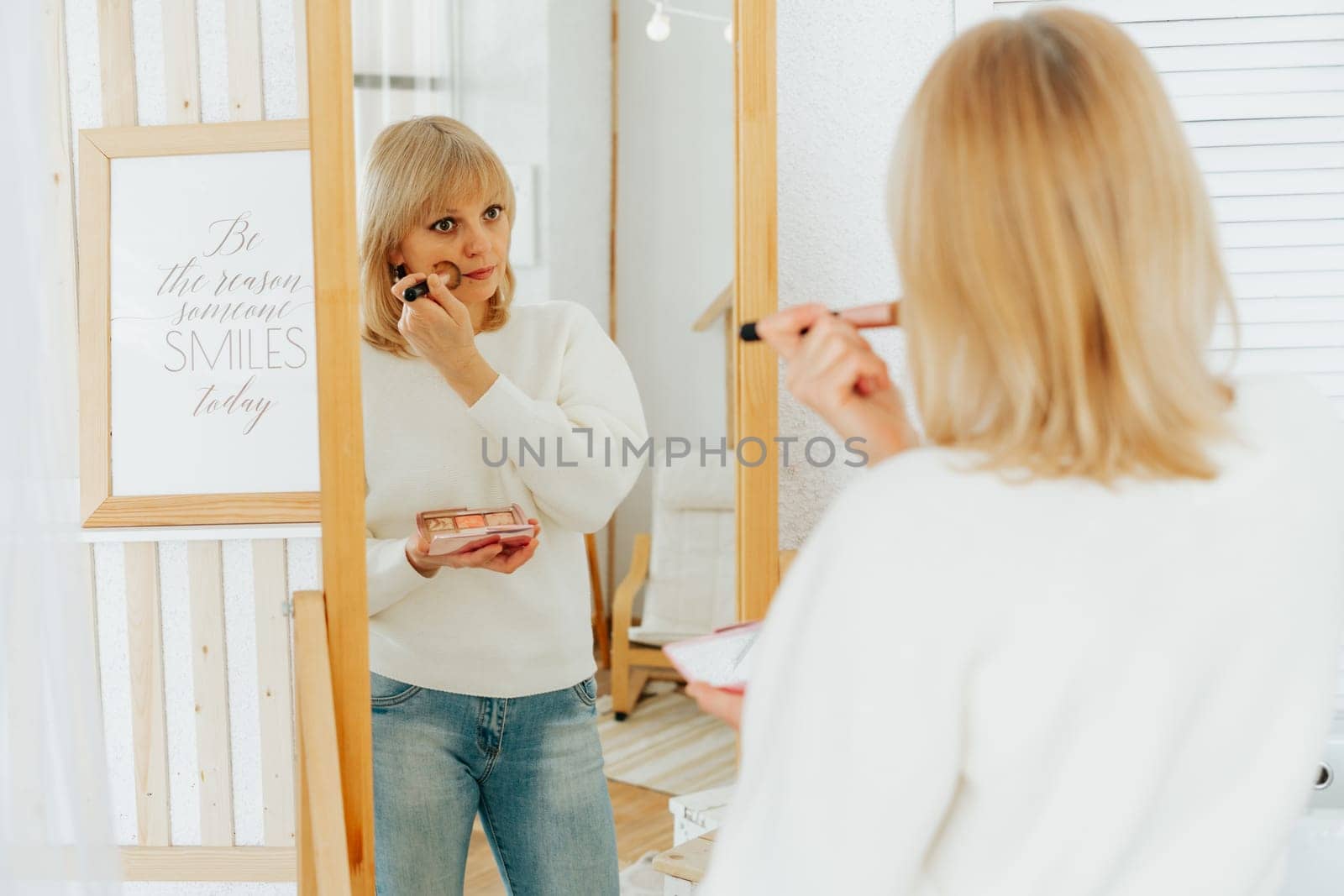 Beautiful smiling mature senior caucasian woman with blonde hair standing near mirror with make up palette and brush, doing makeup and looking at reflection in living room at home.