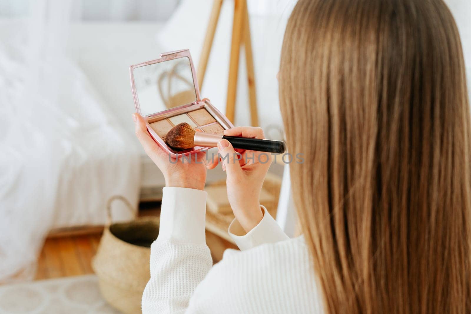 Close up of cropped caucasian woman with long hair holding powder palette and brush, doing makeup and looking at reflection in mirror at home.