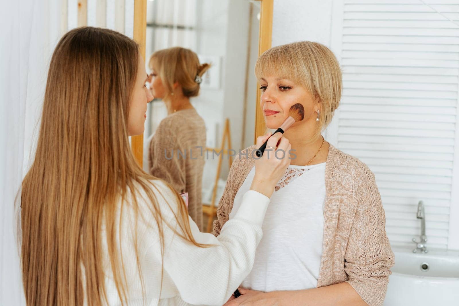 Beautiful smiling young caucasian mature senior mother mom and adult daughter doing makeup, standing near mirror, and looking at reflection in living room home. Family with two women relax together.