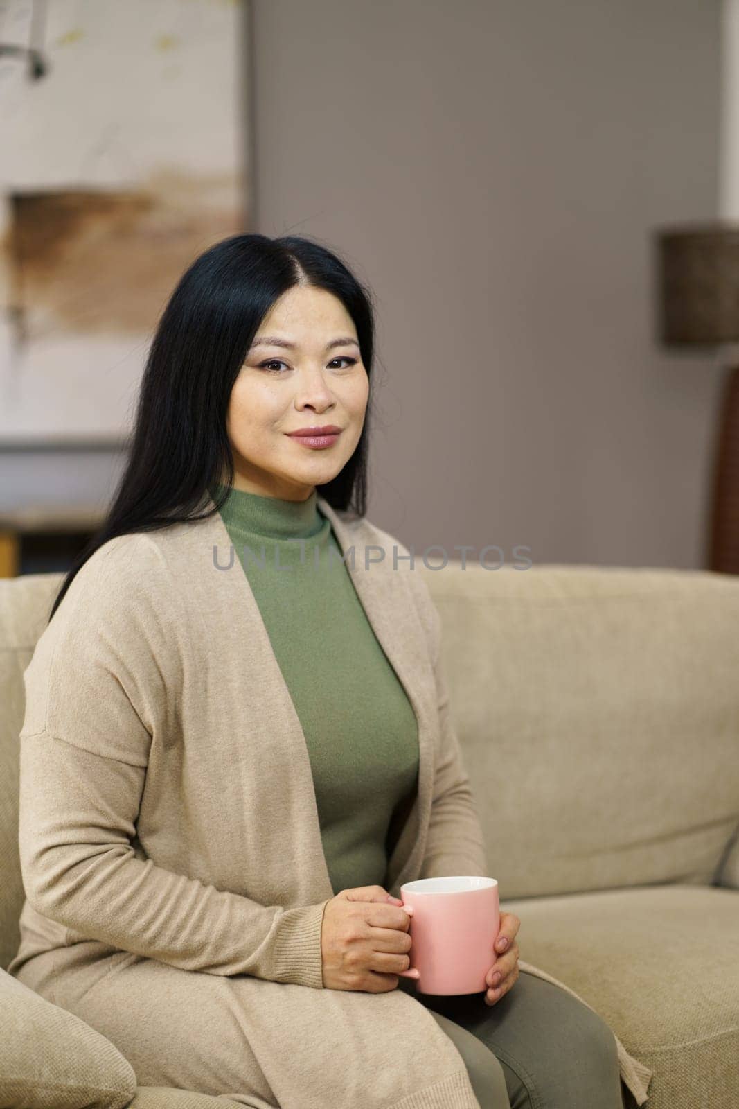 Cute Asian MILF sitting on couch with cup of coffee home. The image captures moment of relaxation and leisure as woman enjoys her coffee break in comfort of her own home. . High quality photo