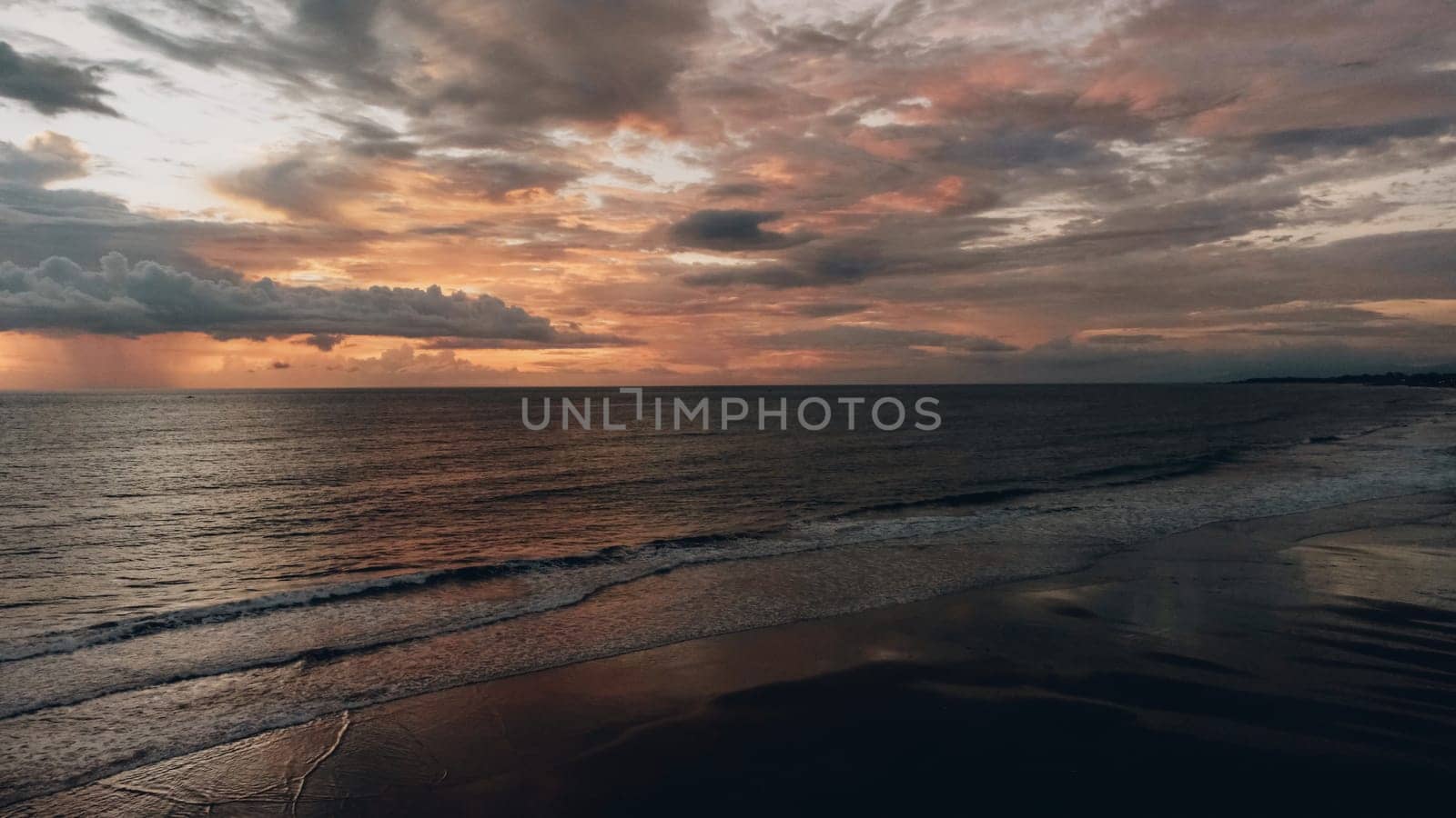 Aerial view of beautiful sunset at the beach. The sky is a mix of grey, black, and orange-red. Waves crash against the shore, reflecting the light