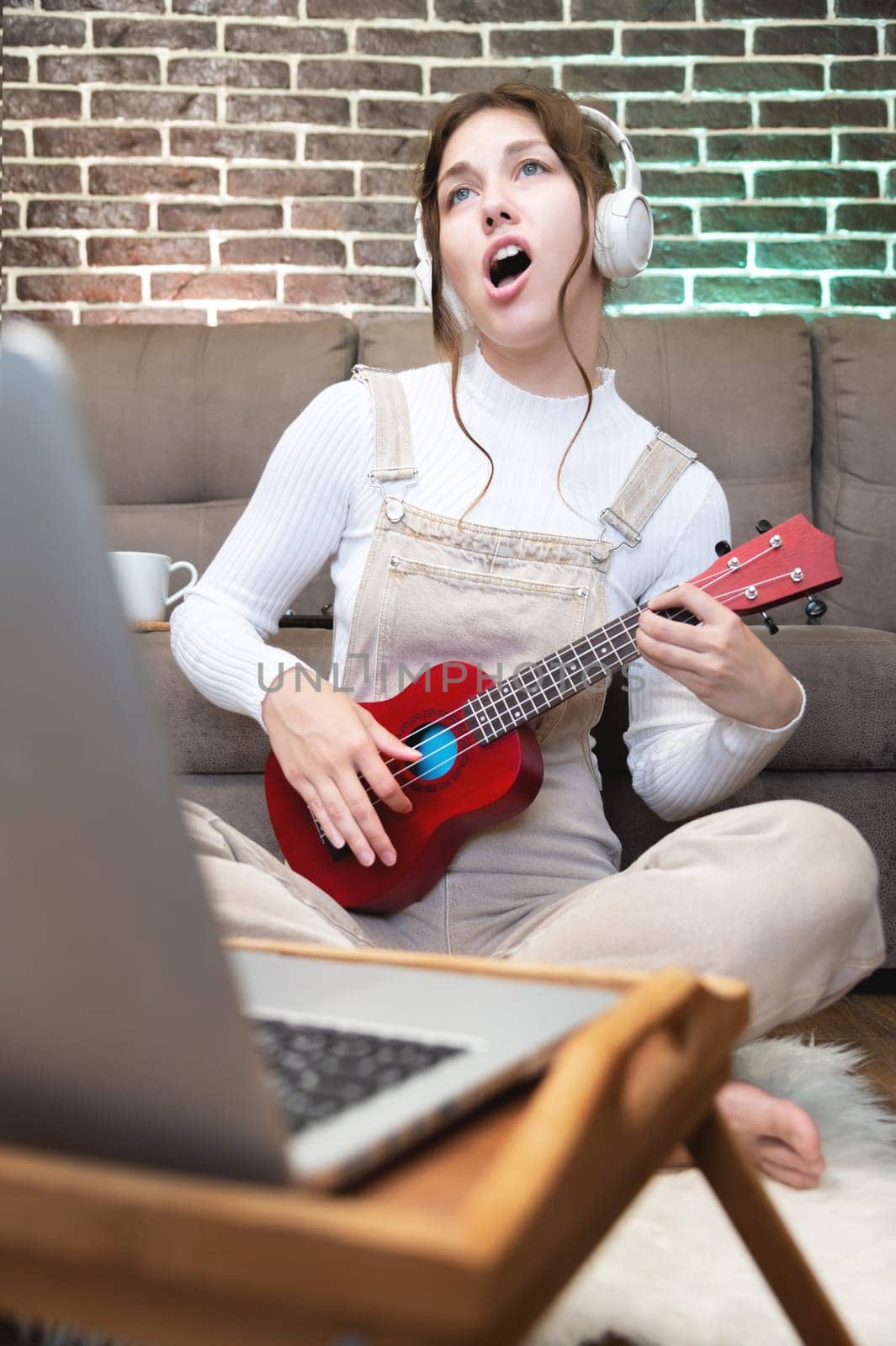 An attractive Caucasian young woman in headphones with a ukulele in her hands sings expressively at home next to a sofa and a laptop. Learning to play stringed musical instruments.
