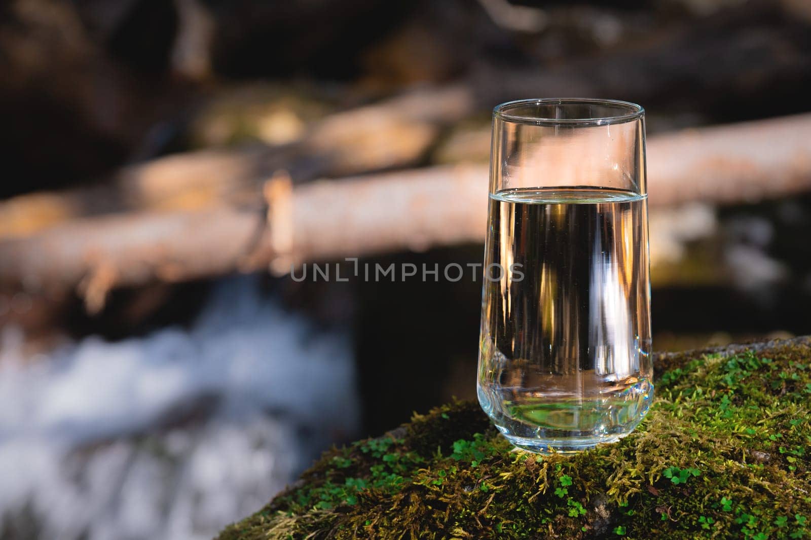Purified fresh drinking water in a glass goblet from a mountain lake stands in the sunlight on a stone covered with moss, against the backdrop of a waterfall.