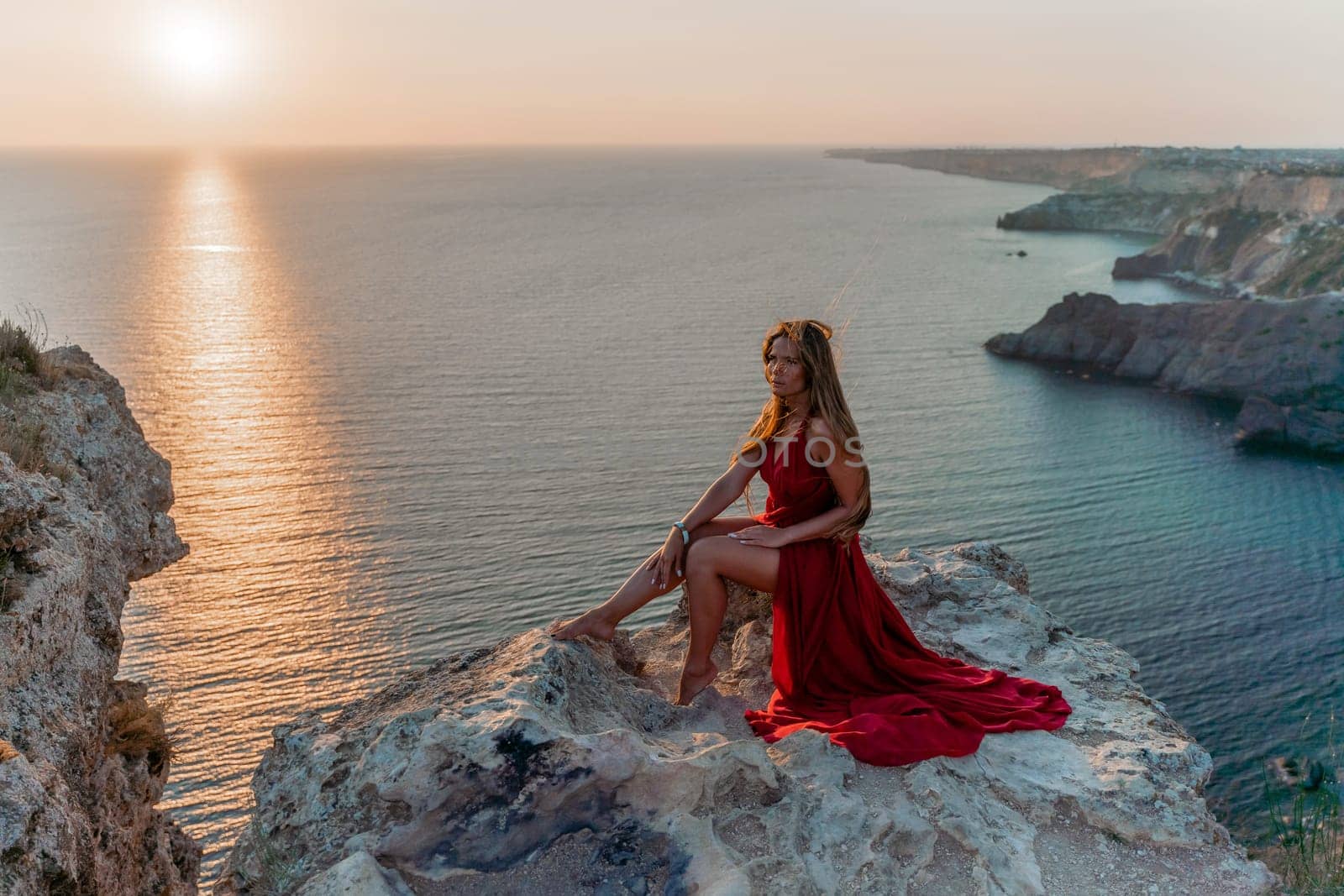Woman sunset sea red dress, side view a happy beautiful sensual woman in a red long dress posing on a rock high above the sea on sunset