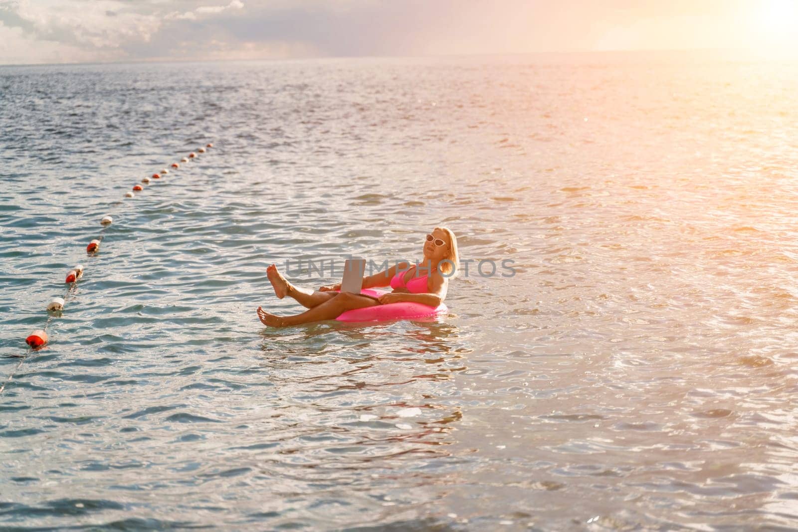 Woman works on laptop in sea. Freelancer, blond woman in sunglases floating on an inflatable big pink donut with a laptop in the sea at sunset. People summer vacation rest lifestyle concept