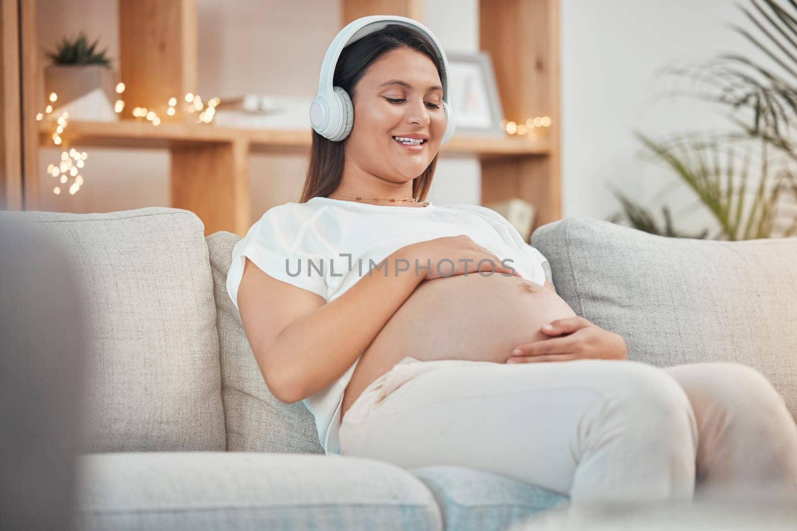 Pregnant, music and relax with a woman sitting on a sofa in their living room of her home expecting a baby. Pregnancy, wellness and stomach with a young female streaming audio alone in her apartment by YuriArcurs