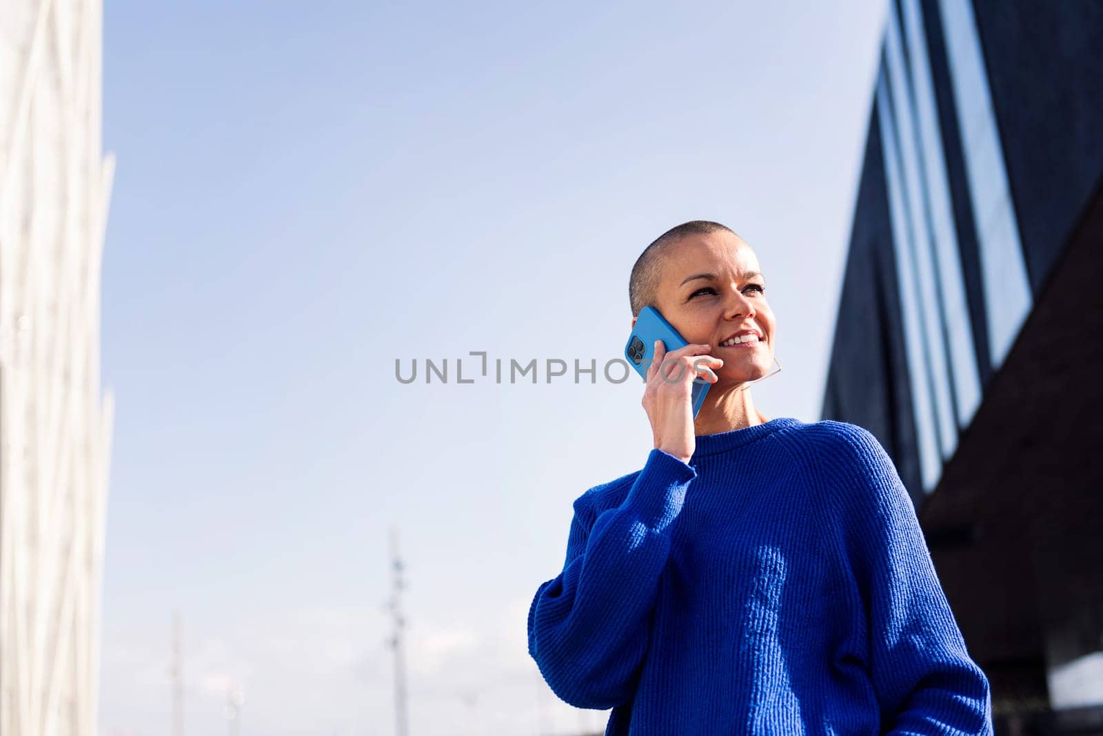 confident young woman with buzz cut in blue sweater talking happy on mobile phone at city, concept or modern lifestyle and technology of communication, copy space for text