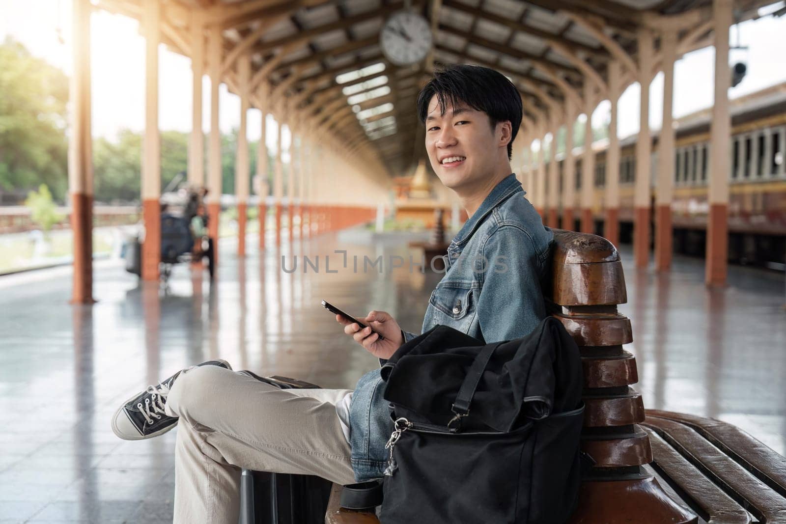 Traveler young asian man using cellphone booking trip at train station. Happy tourist travel by train using smartphone searching location. Male Backpacker arrival at platform railway.