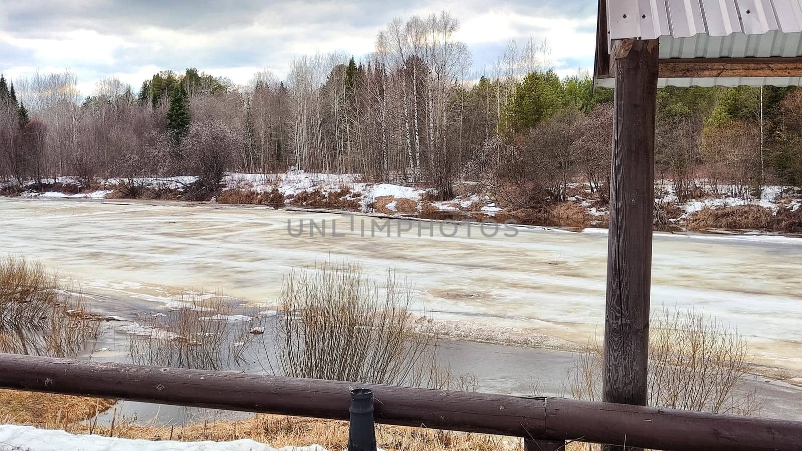 Small river with water under wet swollen ice ready for ice drift, sky with low dark clouds and forest in background in early spring afternoon. Nature landscape during trip to the rural countryside