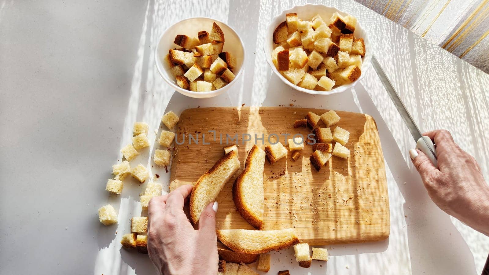 Homemade cooking and cutting of slices of diced dry white rye bread on cutting board and white plate. Hand of woman with Crackers for food, healthy eating on table in sunny day. Top view