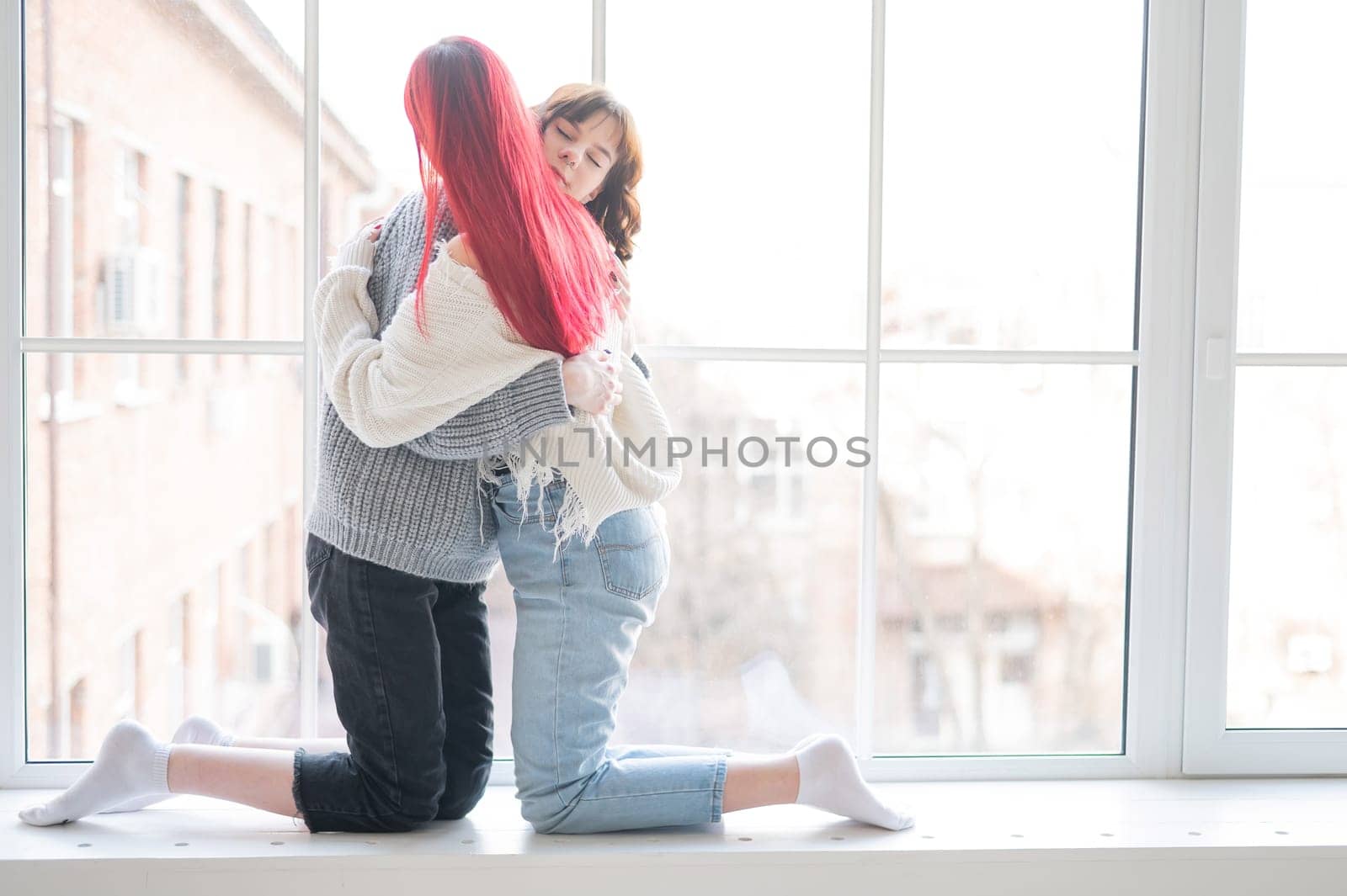 Two women dressed in sweaters sit by the window and gently hug. Lesbian intimacy. by mrwed54