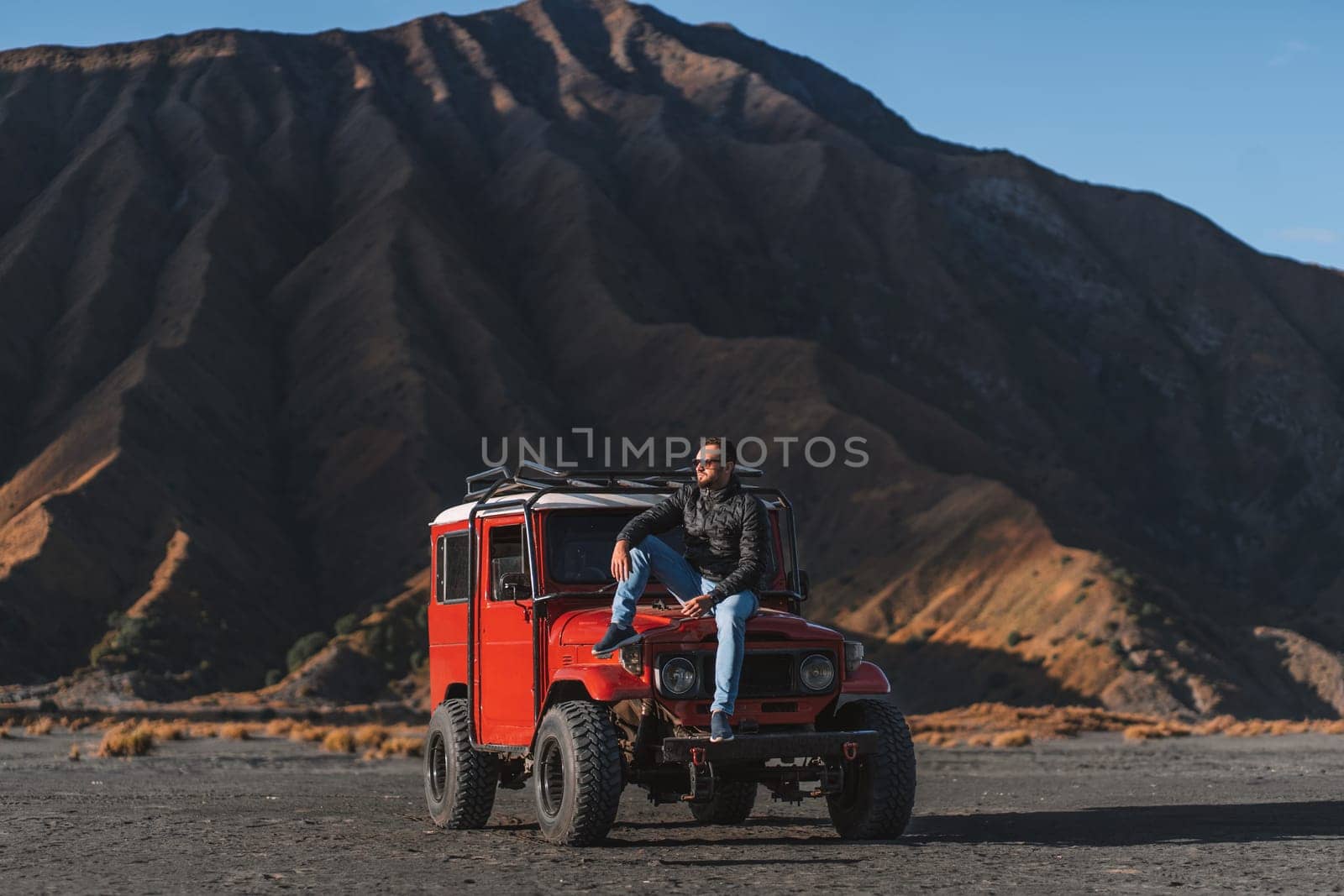 Cool guy sitting on red offroad jeep with Bromo mountain background. Tourist visiting Semeru national park
