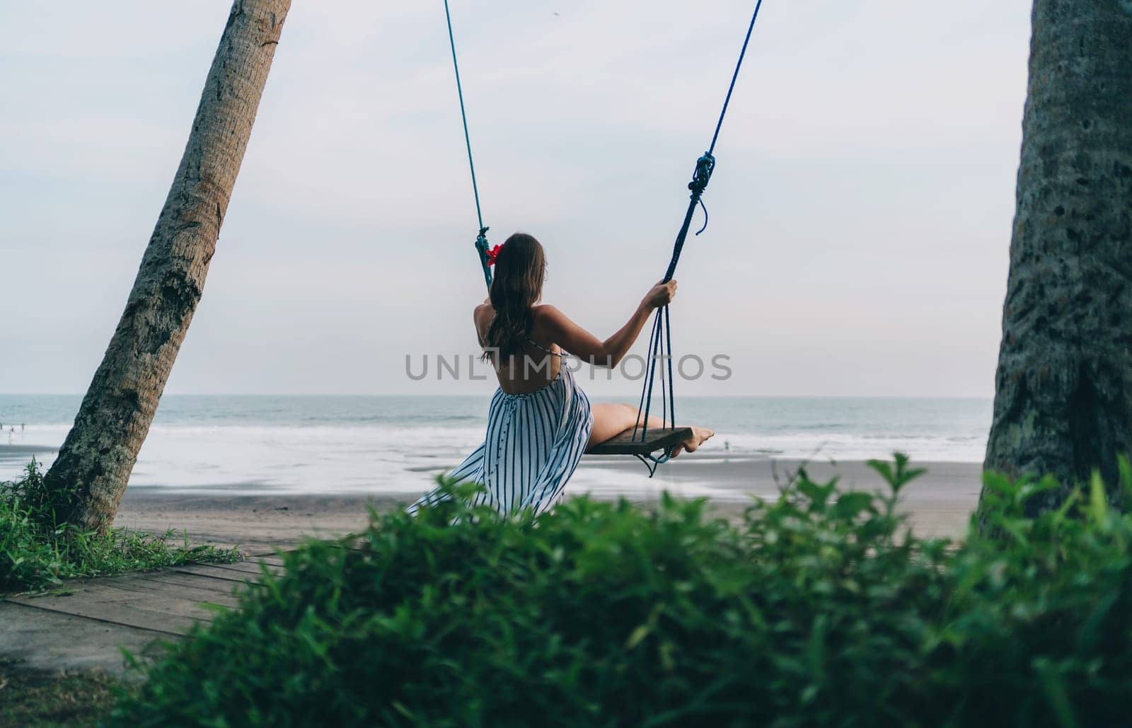 Back view of girl swinging on a swing with wonderful sea view. Young lady on tropical beach swing