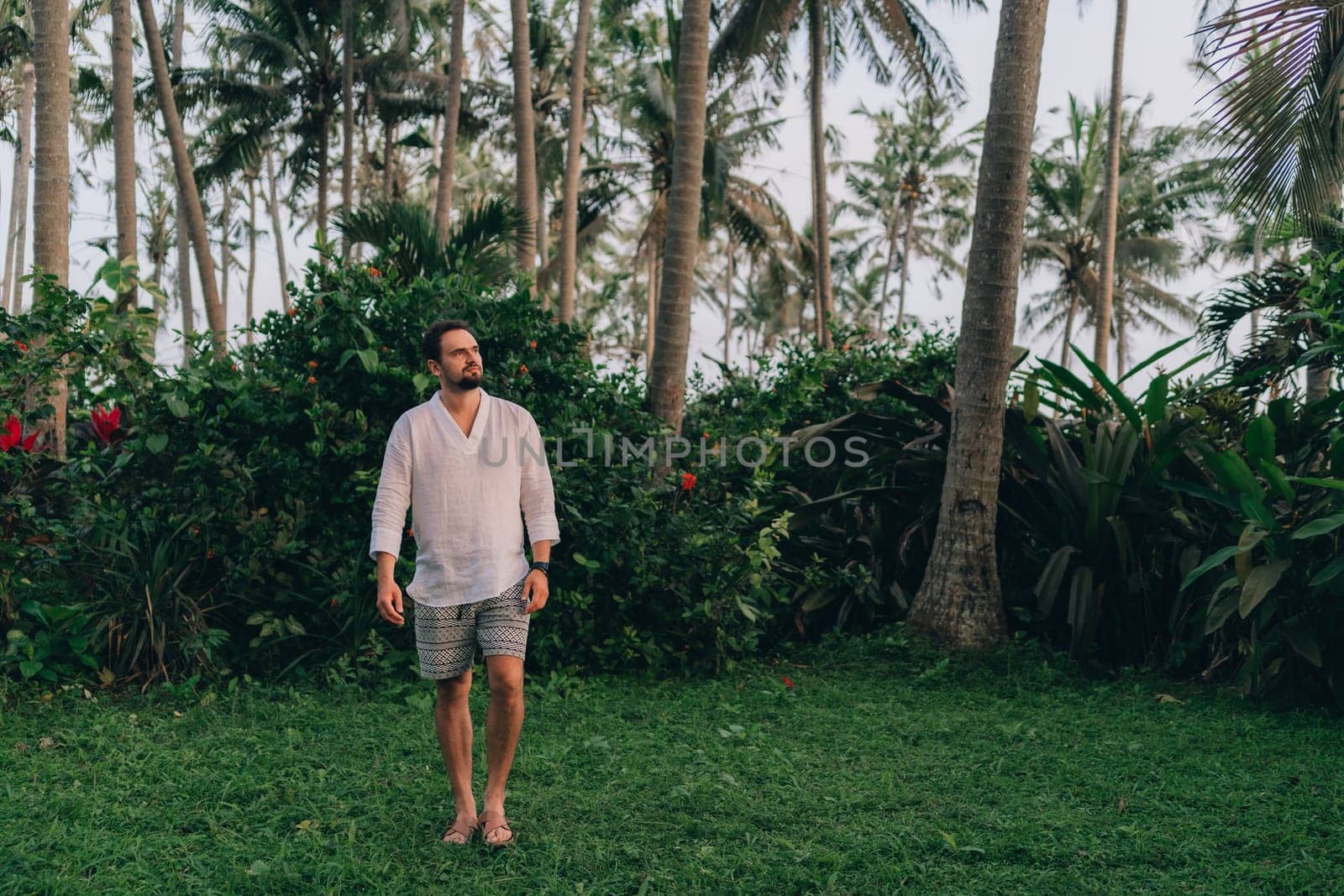 Close up shot of young bearded man in white shirt on green palms background. Male tourist in tropical jungle