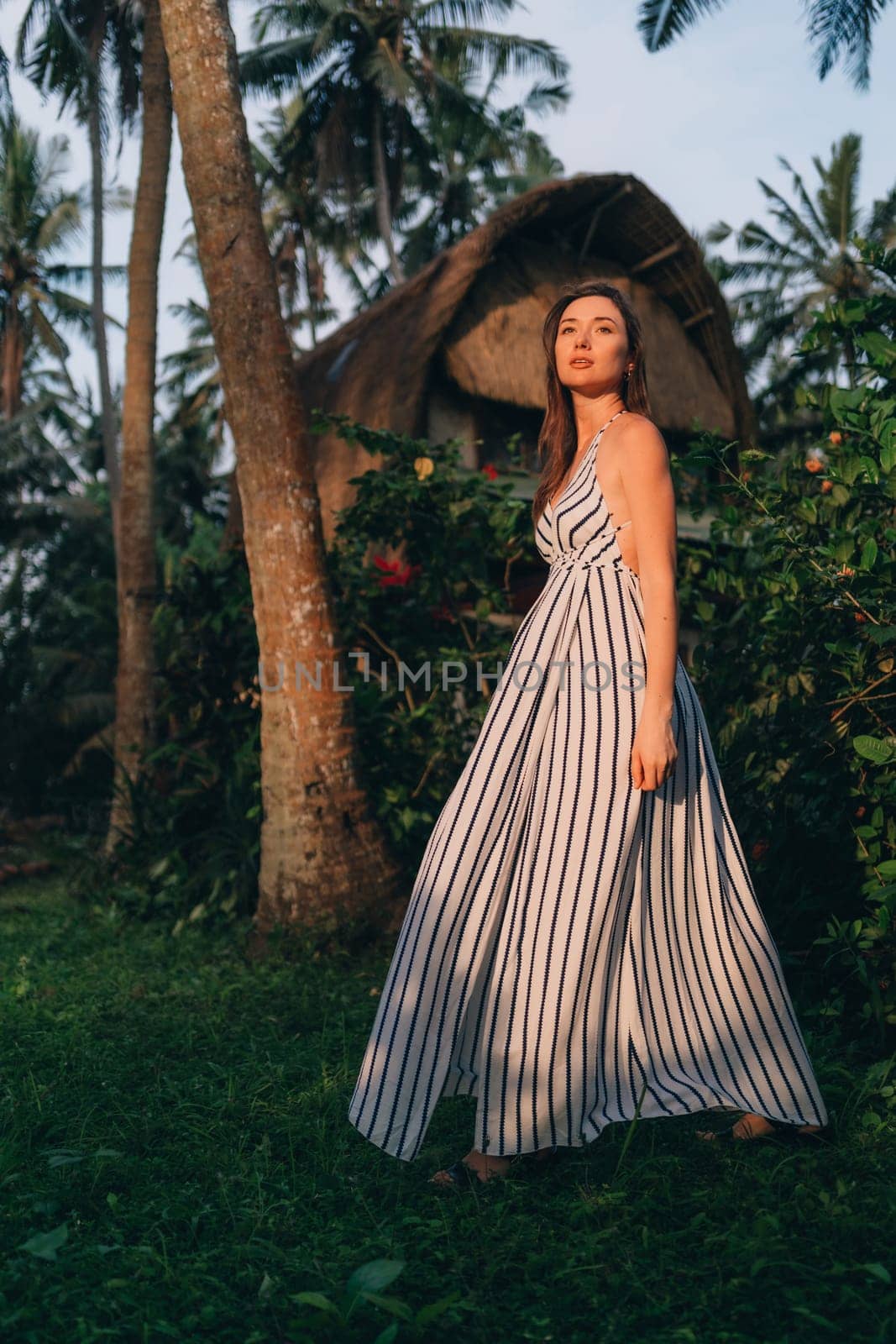 Portrait of beautiful lady in white striped dress with balinese vila background. Young model girl posing in tropical jungle nature