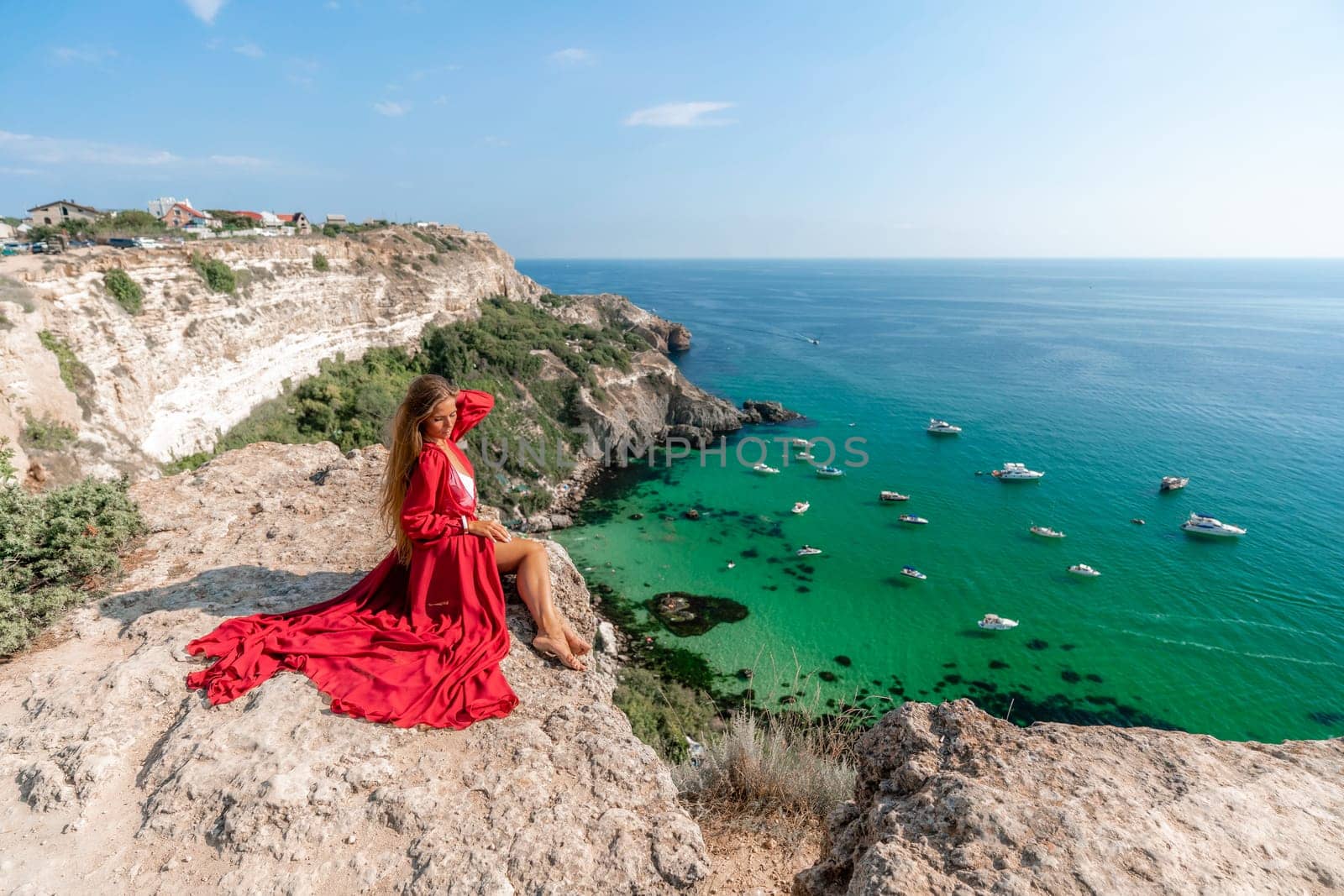 Woman red dress sea. Happy woman in a red dress and white bikini sitting on a rocky outcrop, gazing out at the sea with boats and yachts in the background. by Matiunina