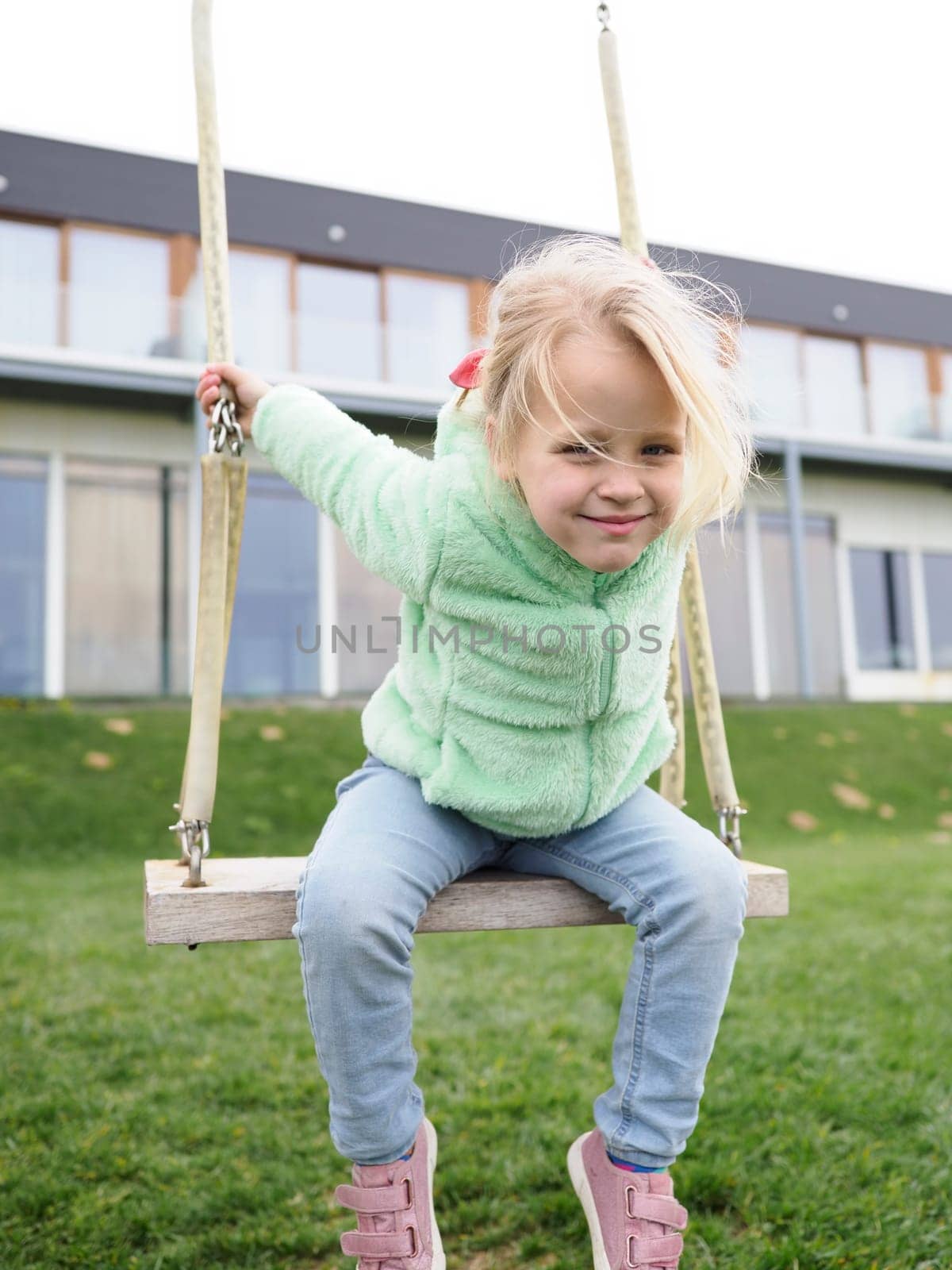 A cheerful little blonde girl, 5 years old, having fun on the swings at the playground near two-storey residential house