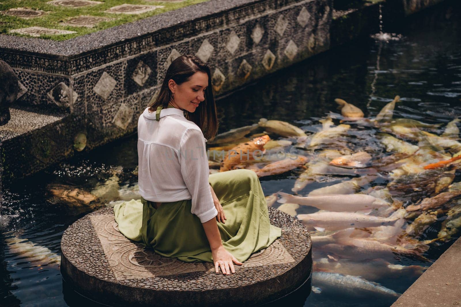 Beautiful girl sitting and admiring pond koi fishes. Smiling girl feeding carps in Ubud Water Palace