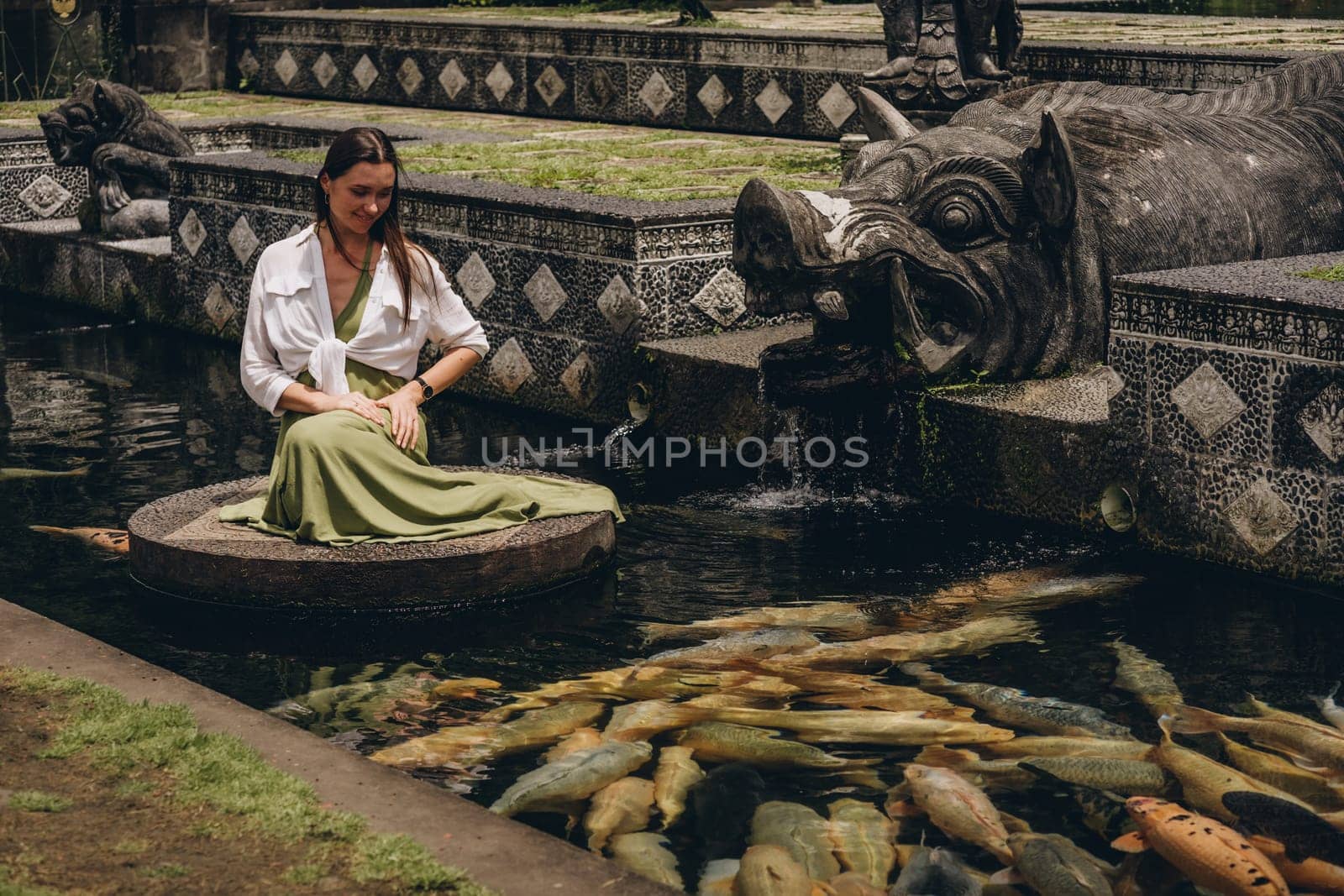 Beautiful girl sitting and admiring pond koi fishes. Smiling girl feeding carps in Ubud Water Palace