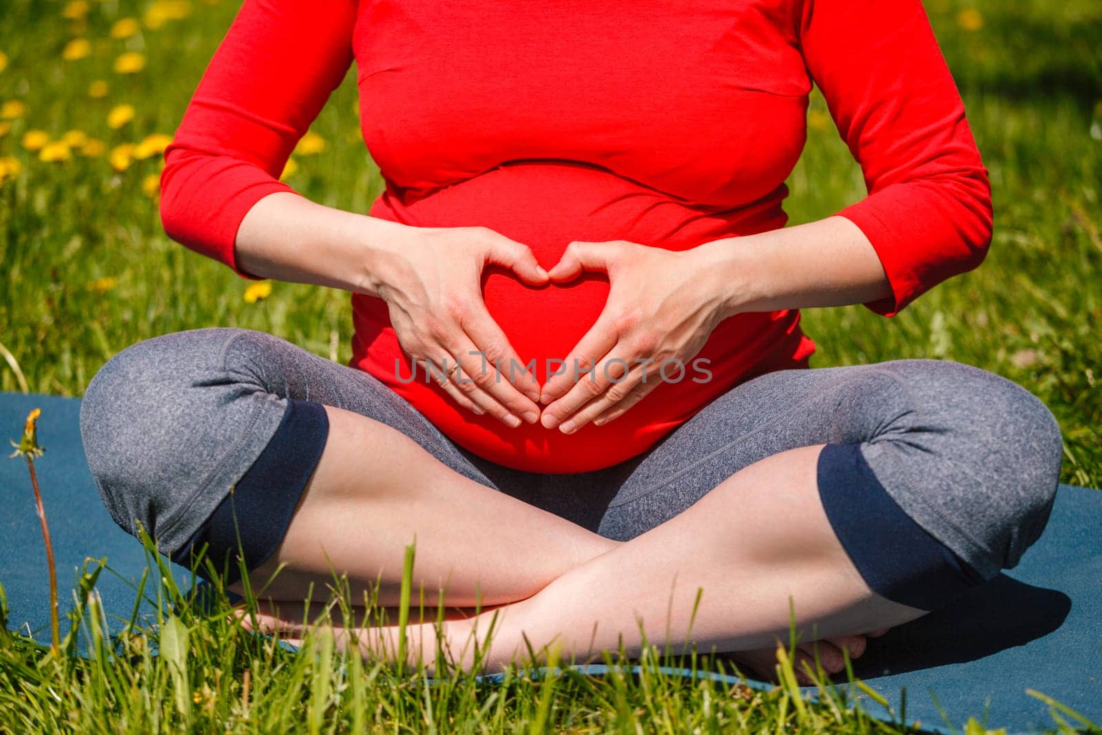 Pregnancy yoga exercise - pregnant woman doing asana Sukhasana easy yoga pose showing heart symbol with hands outdoors on grass lawn with dandelions in summer