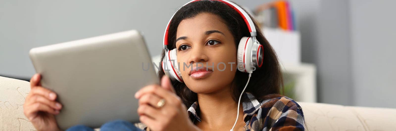 Focused black woman studying foreign language online. Remote female student in virtual classroom sitting on sofa in headset and holding tablet