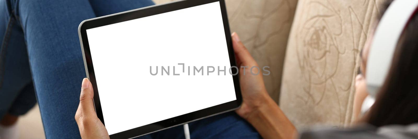 Mockup image of a digital tablet with black screen in hands of woman in headphones. Person resting at home on sofa with tablet over shoulder view
