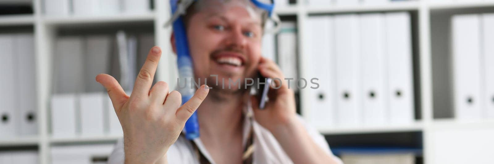 Emotionally cheerful male manager in travel company in swimming goggles is talking on mobile phone at workplace. Sale hot tourist tours travel and vacation concept