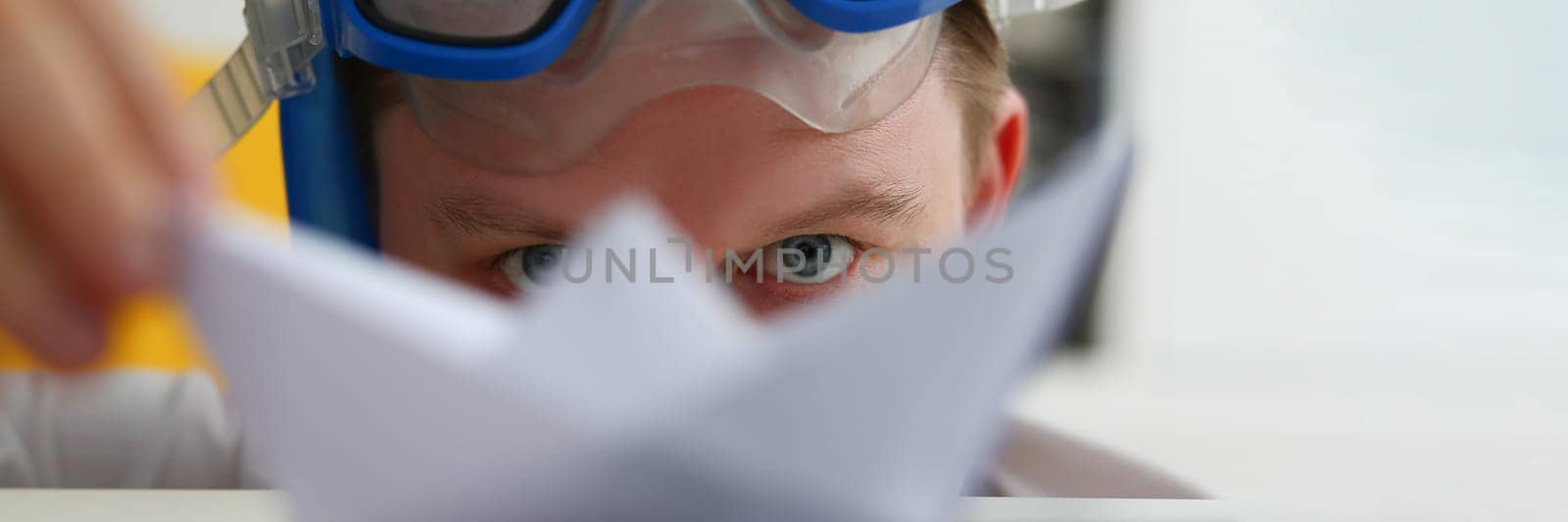 Young man in swimming goggles plays with paper ship at workplace by kuprevich