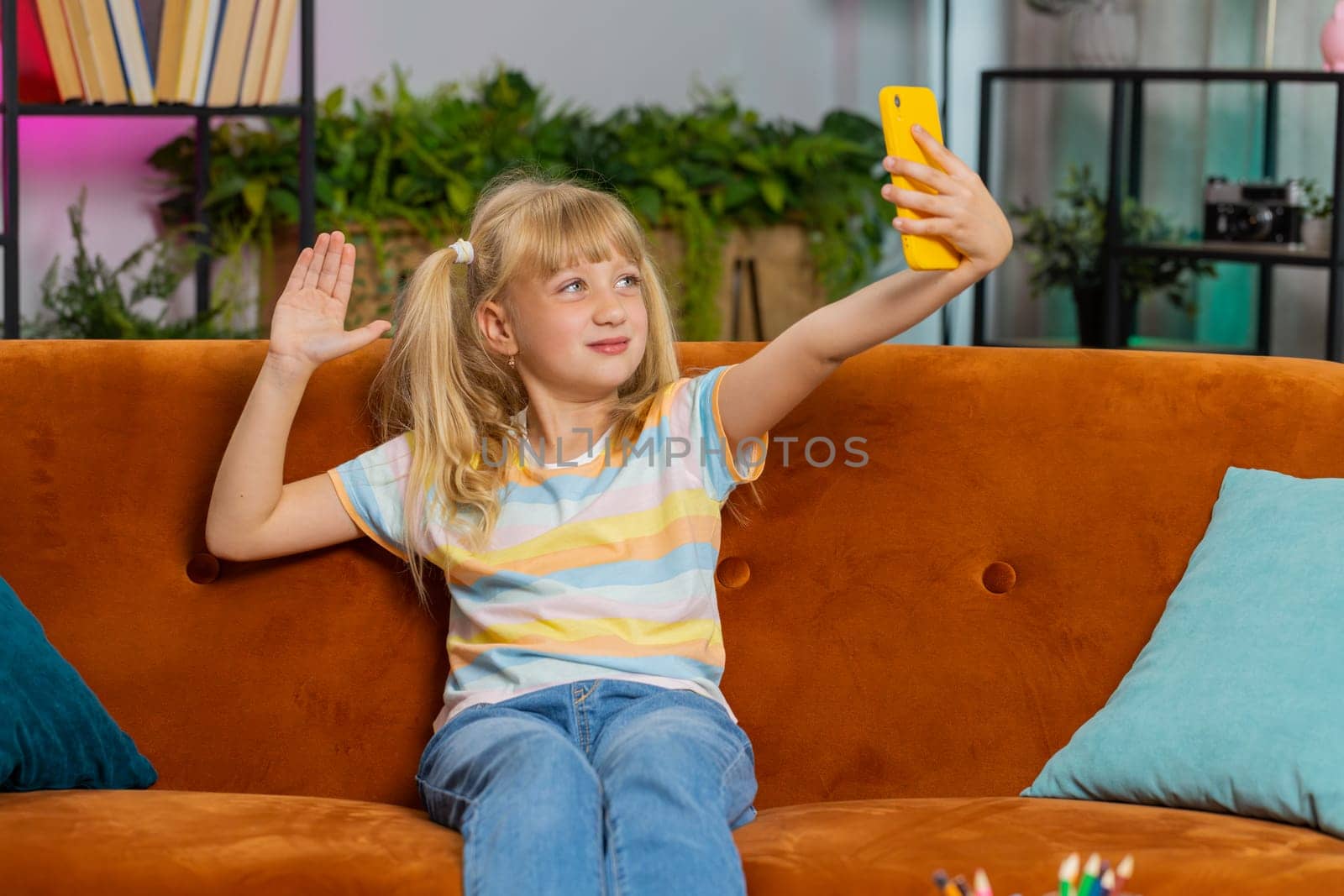 Young blonde child kid girl blogger taking selfie on smartphone, communicating video call online with social media subscribers. Female teen toddler recording video story at home in room sits on sofa