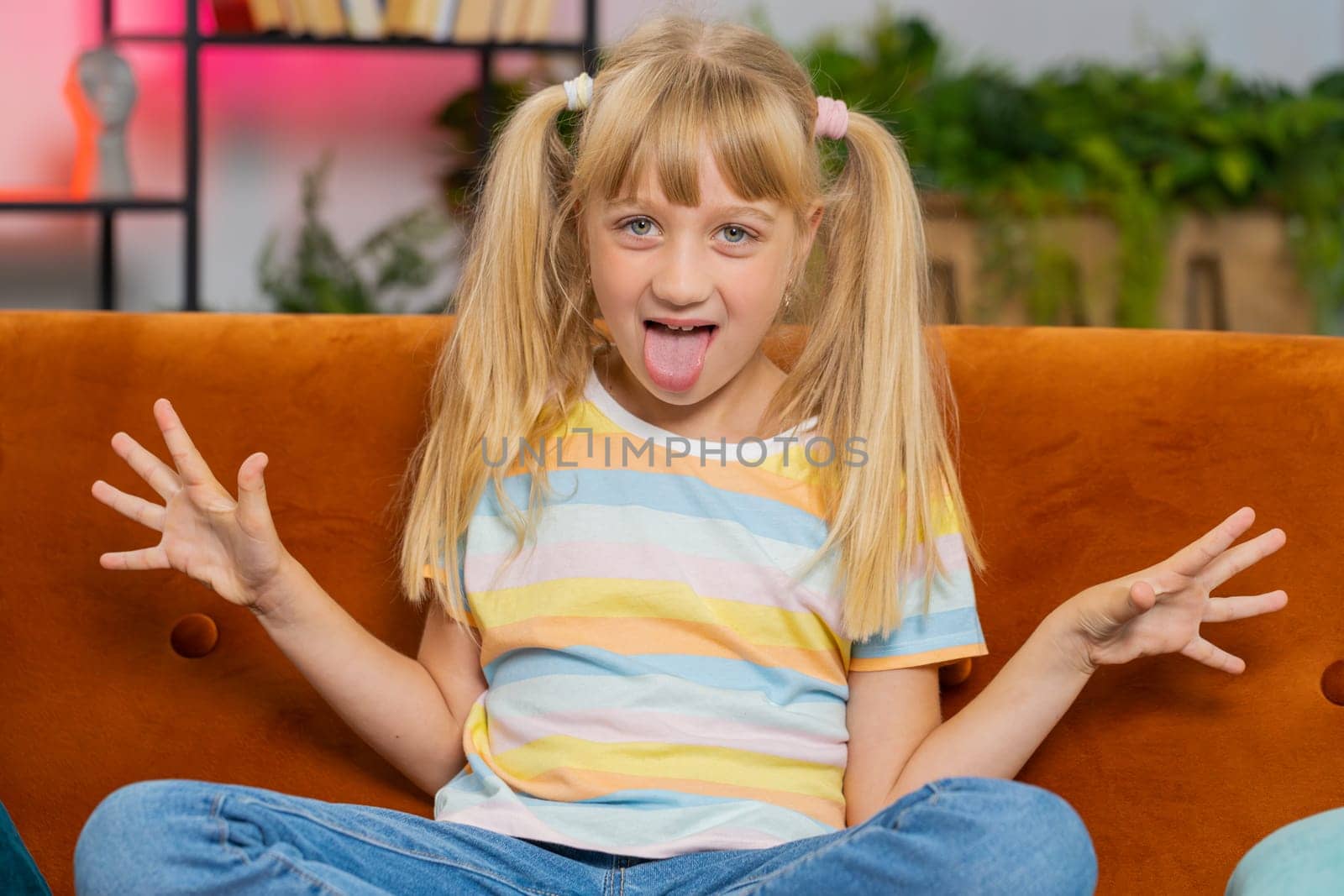 Caucasian joyful preteen school girl making playful silly facial expressions and grimacing, fooling around, showing tongue. Young excited blonde child kid at home room apartment sitting on couch