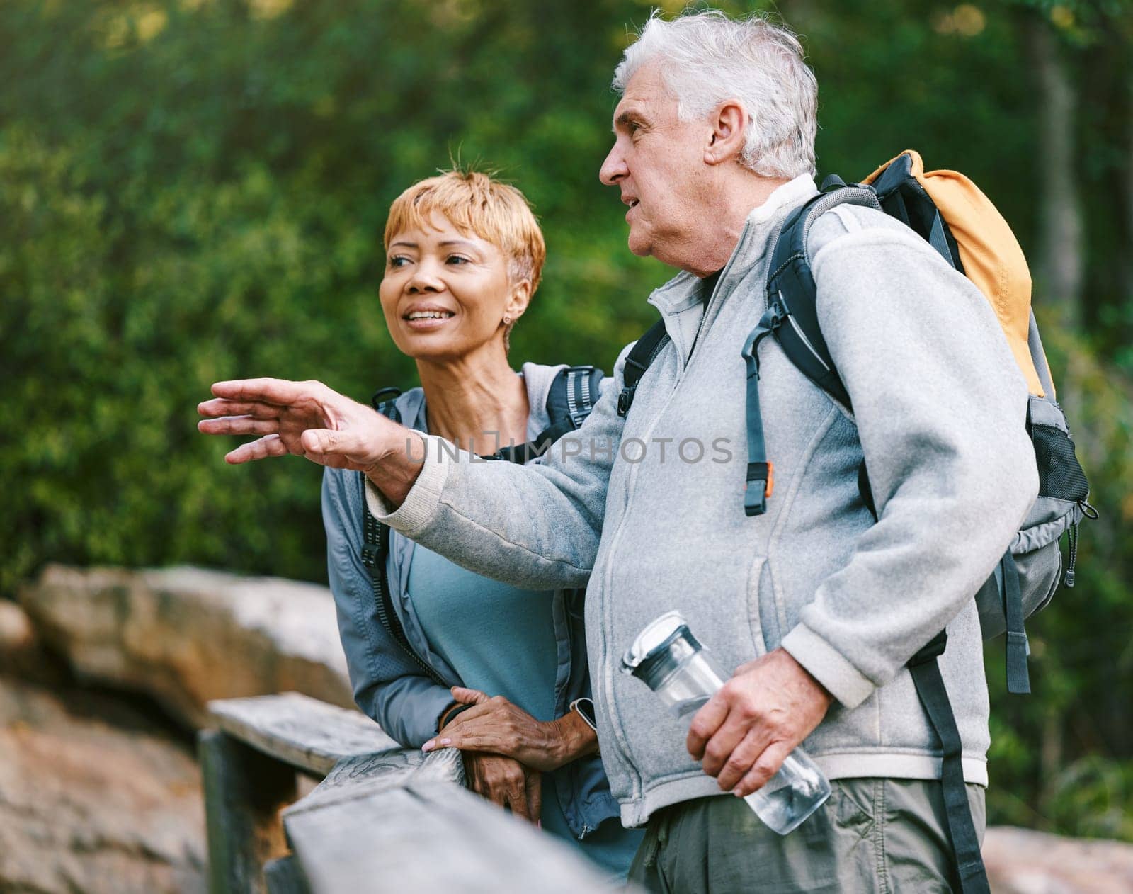 Couple, elderly and hiking in forest with fitness and interracial, talking about view and relax while trekking. Retirement, exercise and hike in nature with water bottle for hydration and travel