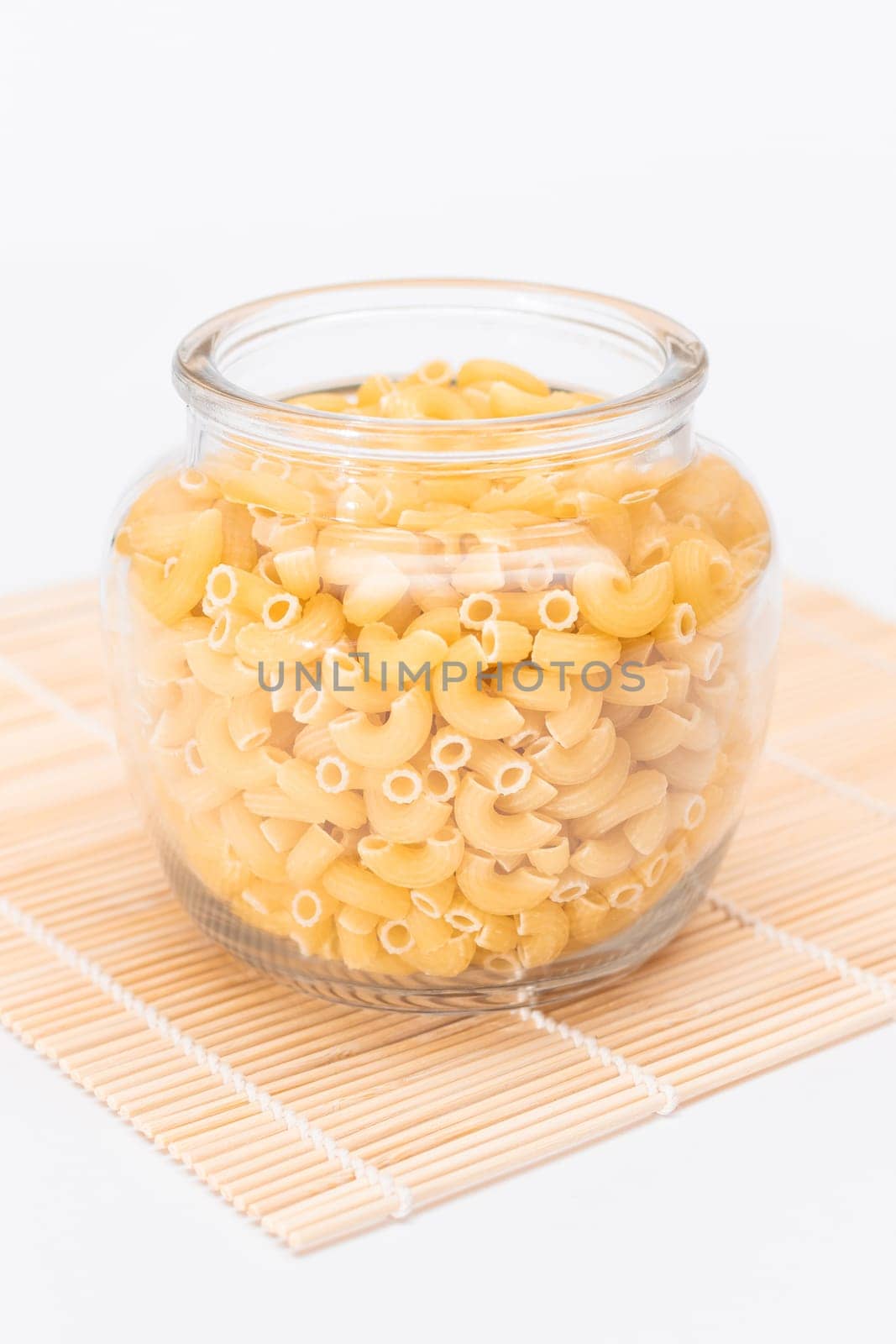 Uncooked Chifferi Rigati Pasta in Glass Jar on Bamboo Mat on White Background. Fat and Unhealthy Food. Classic Dry Macaroni. Italian Culture and Cuisine. Raw Pasta