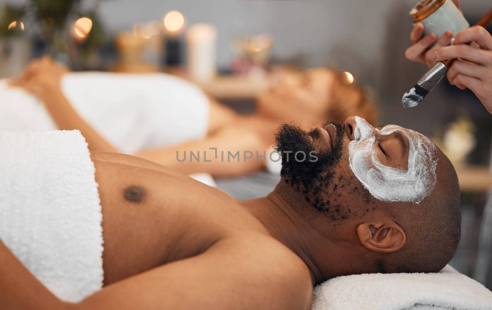 Skincare, wellness and black man getting a facial at a spa for relaxation, calm and satisfaction. Beauty, luxury treatment and couple massage at beauty salon together, skincare products on mans face by YuriArcurs