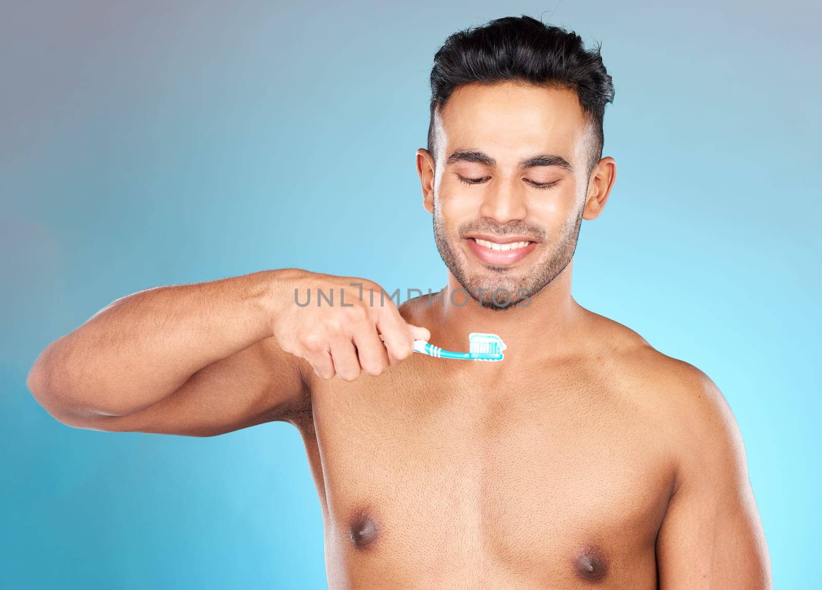 Teeth, dental care and man brushing teeth with toothbrush and toothpaste on blue background with smile on face. Morning routine, healthcare and fresh, happy male model from India in studio portrait