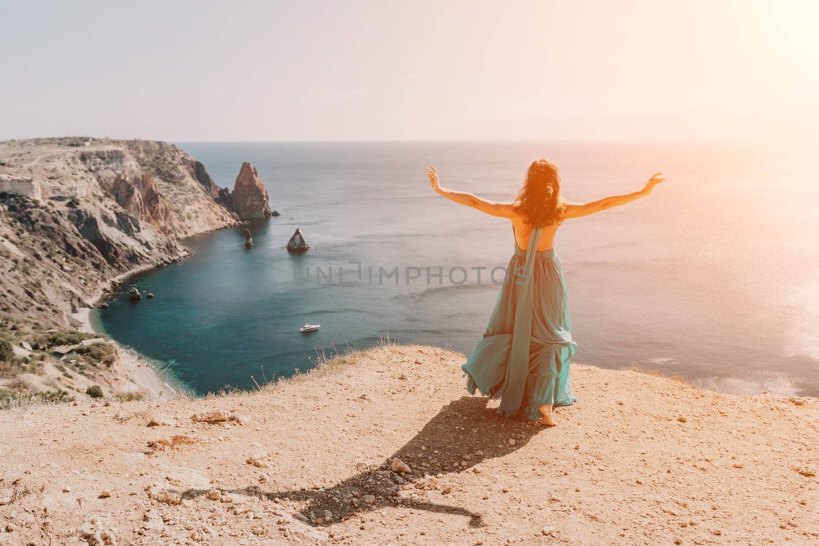 Side view a Young beautiful sensual woman in a red long dress posing on a rock high above the sea during sunrise. Girl on the nature on blue sky background. Fashion photo.