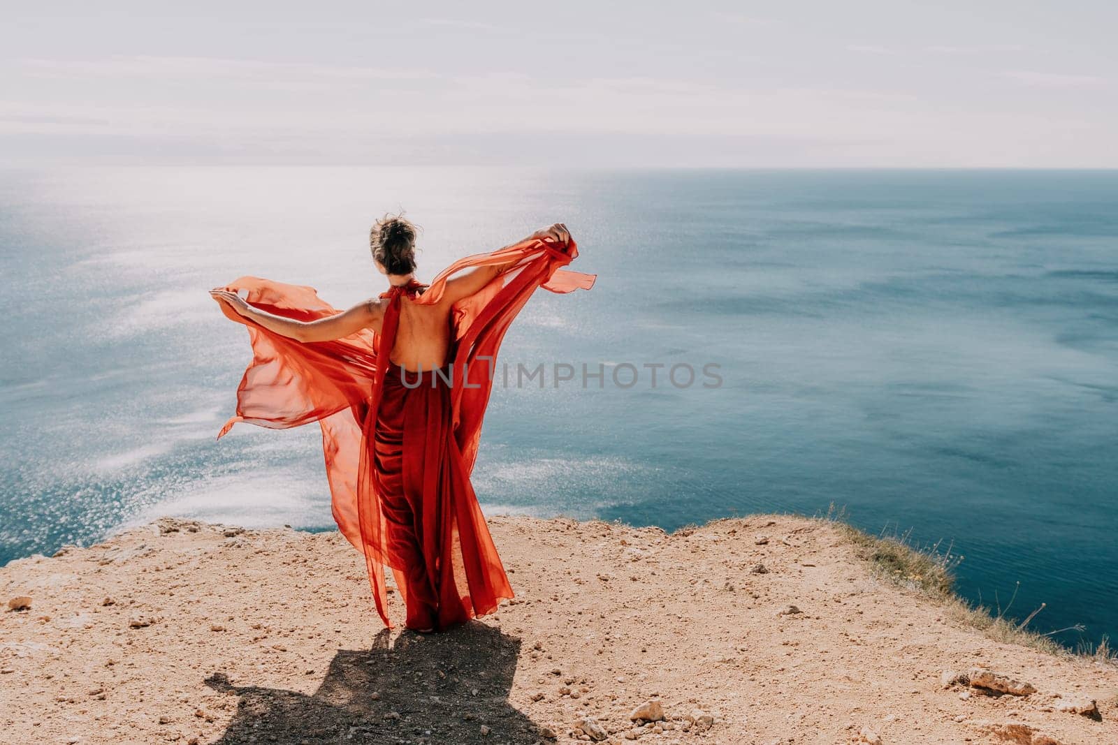 Woman in red dress on sea. Side view a Young beautiful sensual woman in a red long dress posing on a rock high above the sea on sunset. Girl on the nature on blue sky background. Fashion photo. by panophotograph