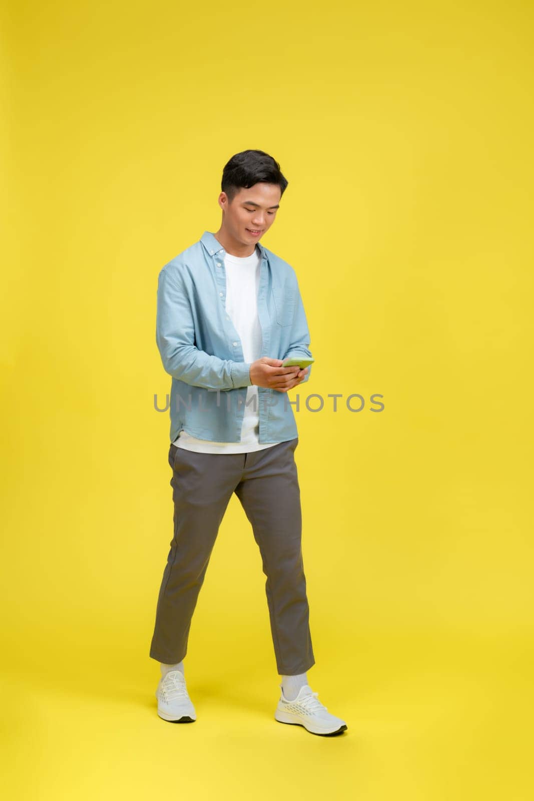 Happy man in casual wear using mobile phone chatting online, walking on yellow background
