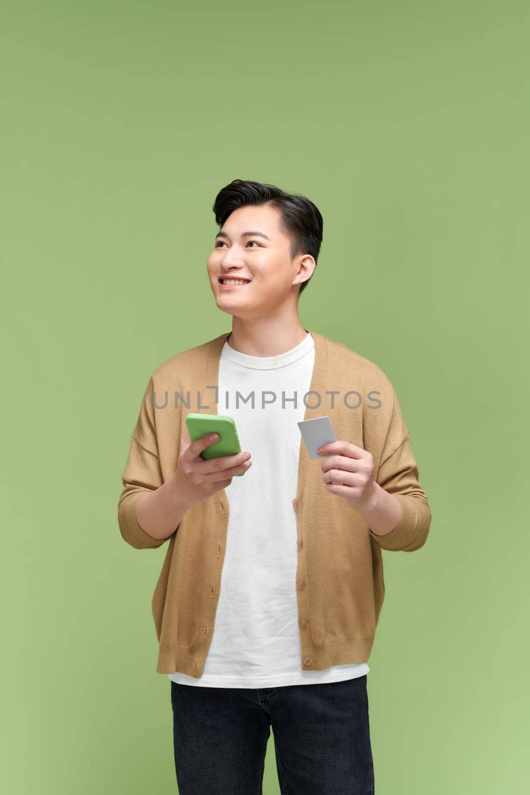 Smiling man holding credit card and mobile phone while standing against green background